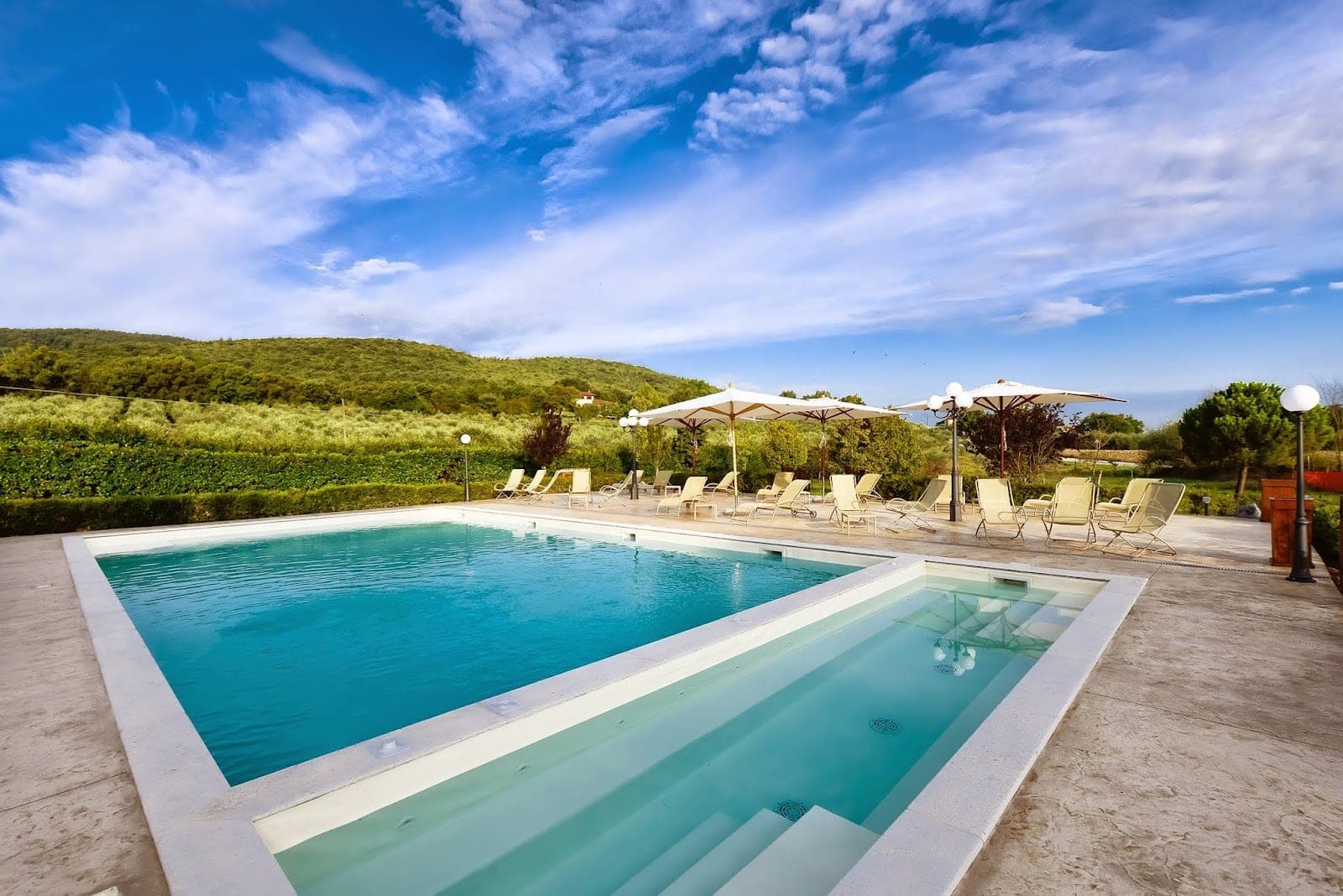 A swimming pool and lounge chairs with a green landscape under a blue sky.