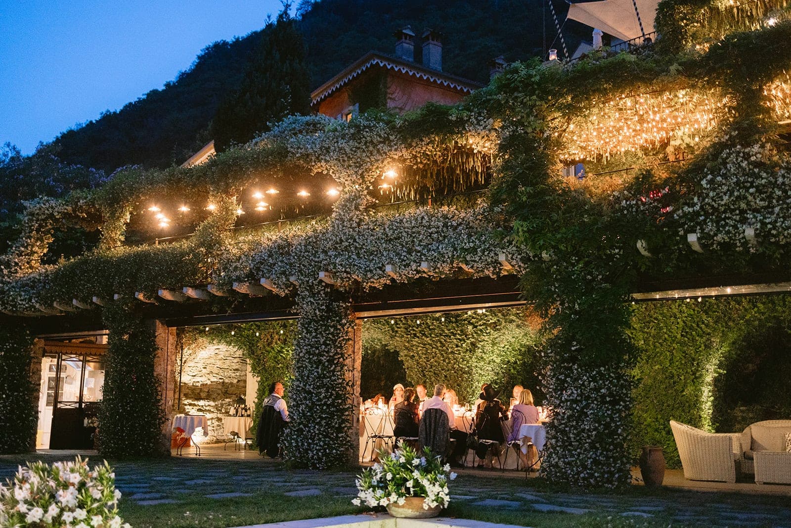 Outdoor reception area with tables set for dinner under a vine-covered pergola with string lights.