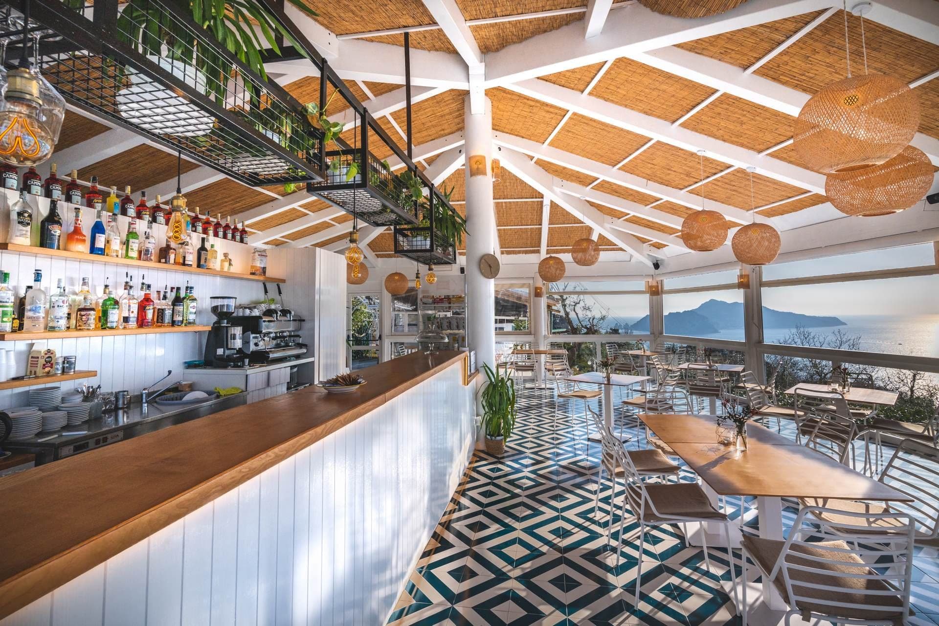 Interior of a restaurant with a bar and tables overlooking the sea.