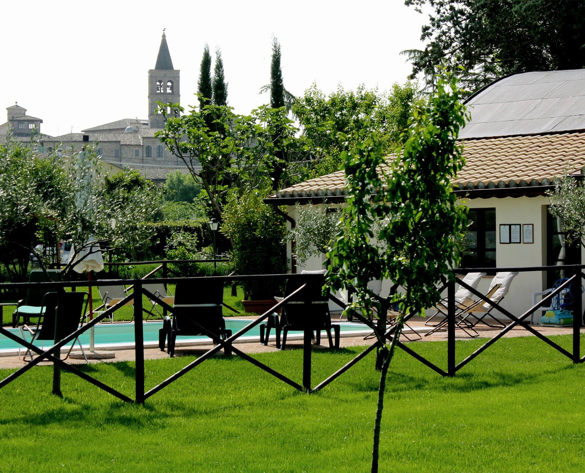 A swimming pool area with lounge chairs and a building, surrounded by greenery and a distant town.