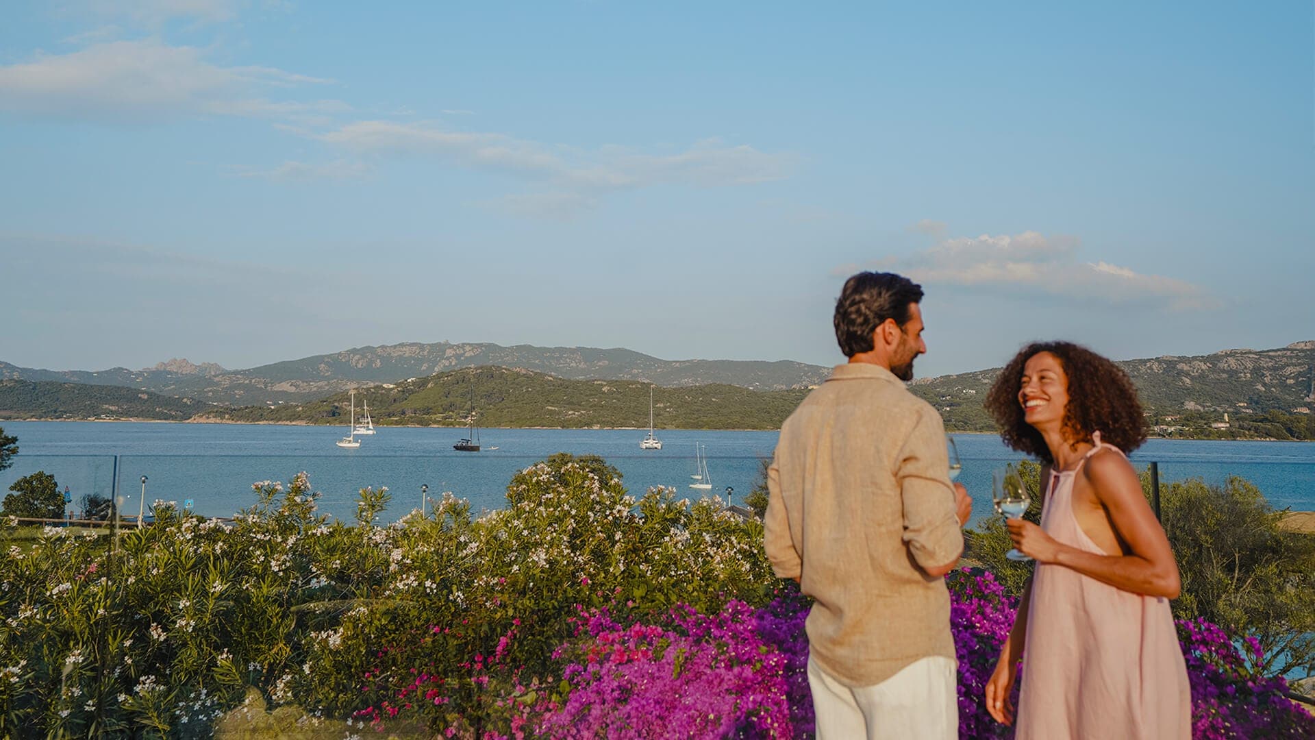 A couple stands on a terrace overlooking a bay with sailboats.