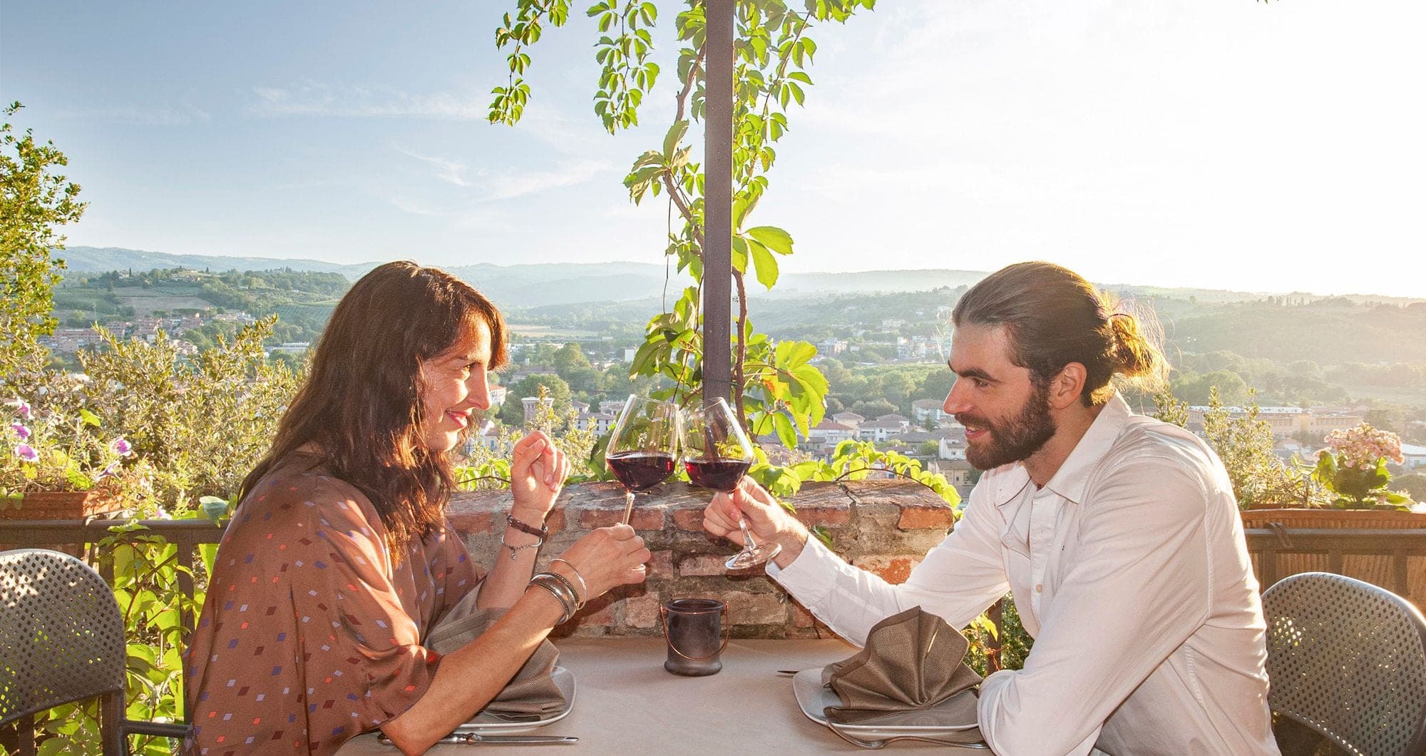 A couple toasting with red wine on a terrace overlooking a valley.