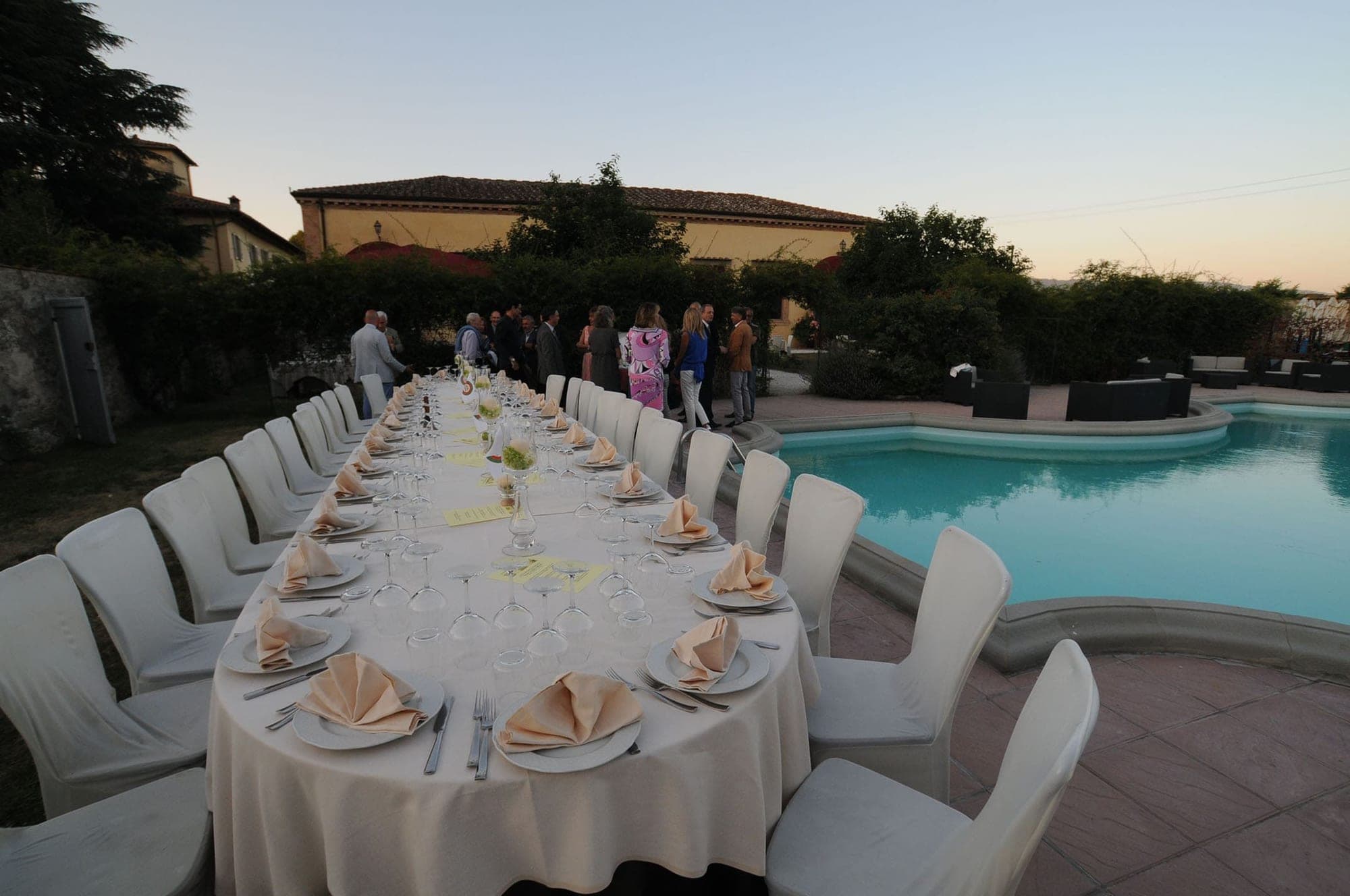 A long reception table is set next to a swimming pool with guests in the background.