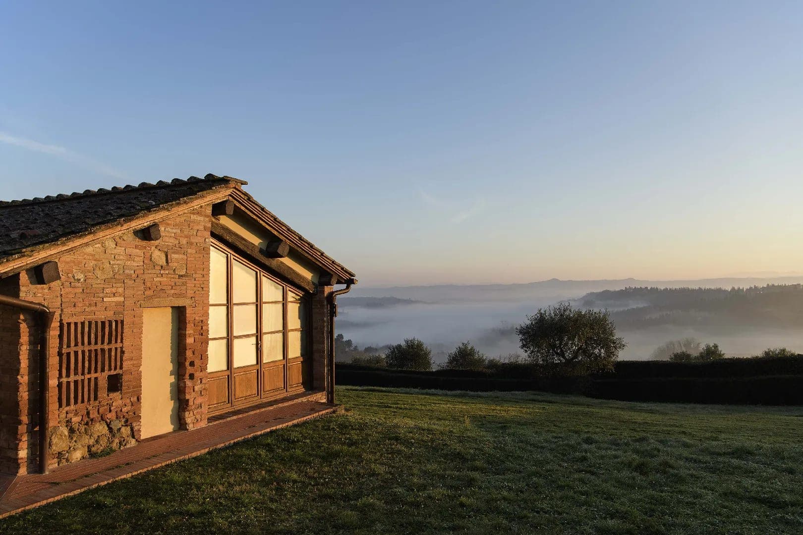 A brick building with large windows overlooks a misty valley at sunrise.