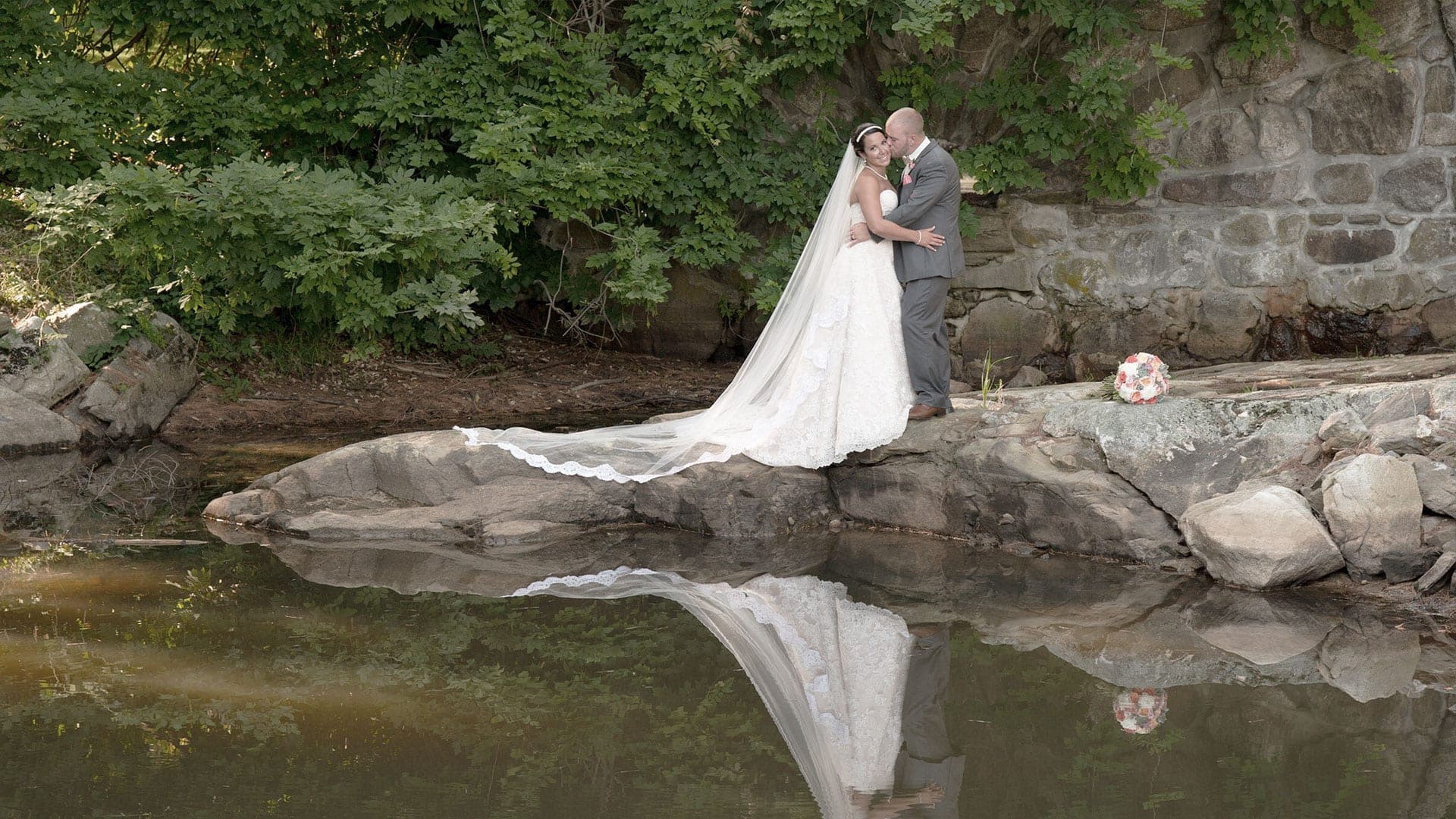 Bride and groom standing on rocks by water with reflection
