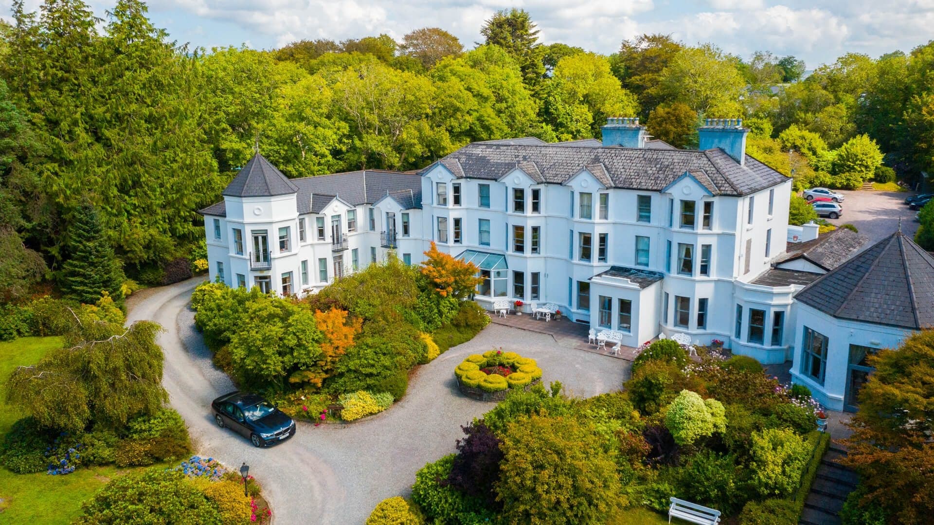 Aerial view of a large white hotel building surrounded by lush green trees and gardens.