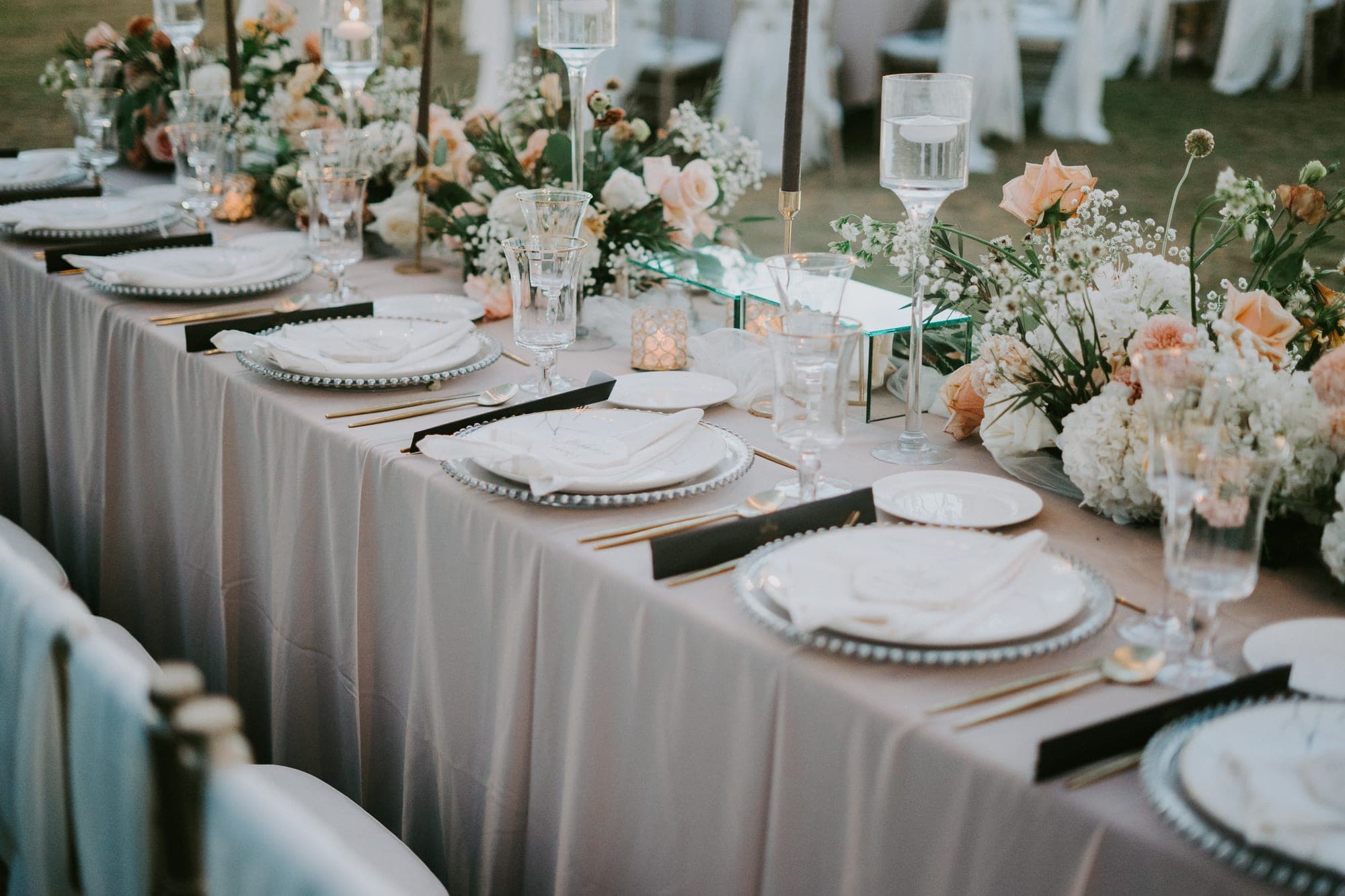 A long reception table is set with white plates, gold cutlery, and floral centerpieces.