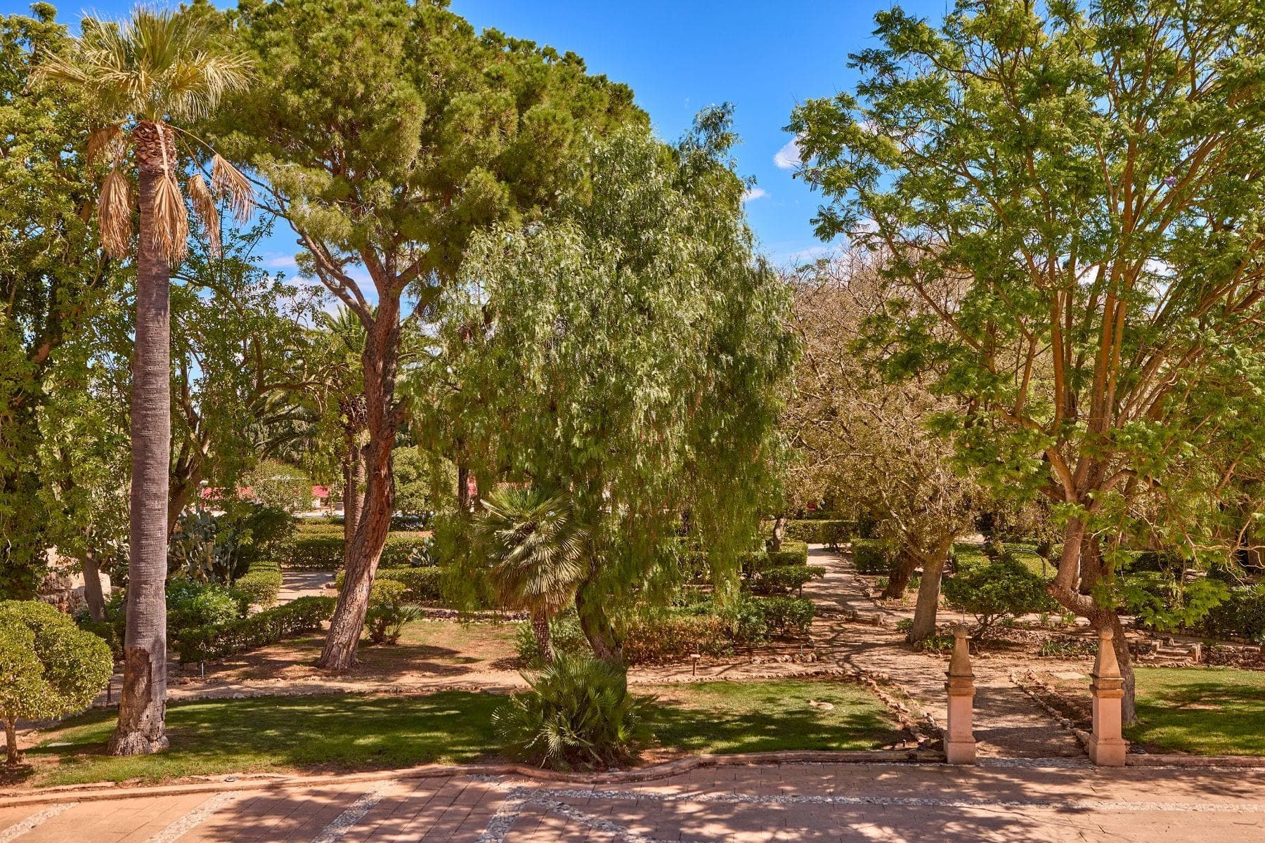 A garden path winds through manicured bushes and trees under a blue sky.