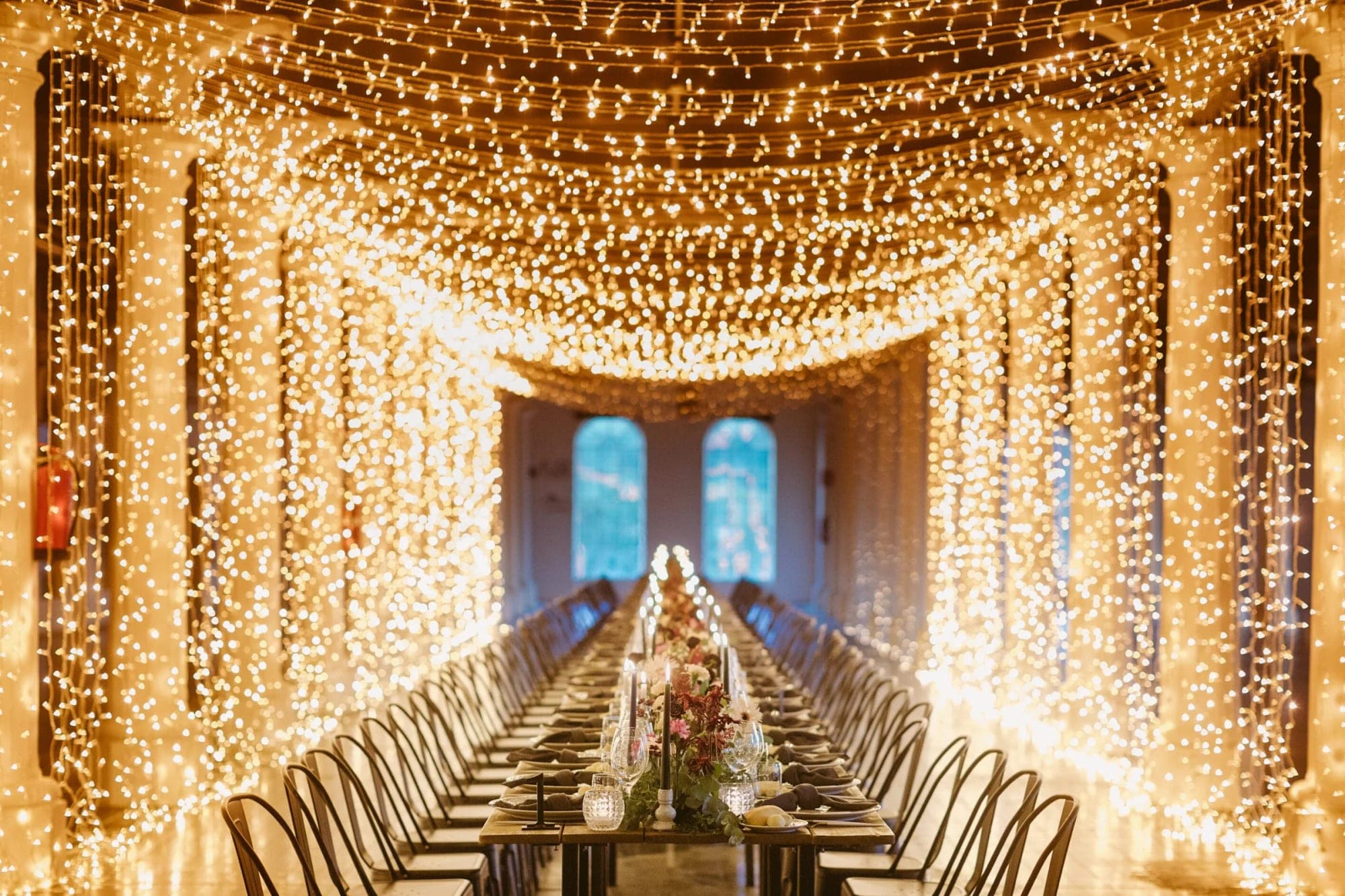 A long banquet table set for a reception, illuminated by strings of fairy lights.
