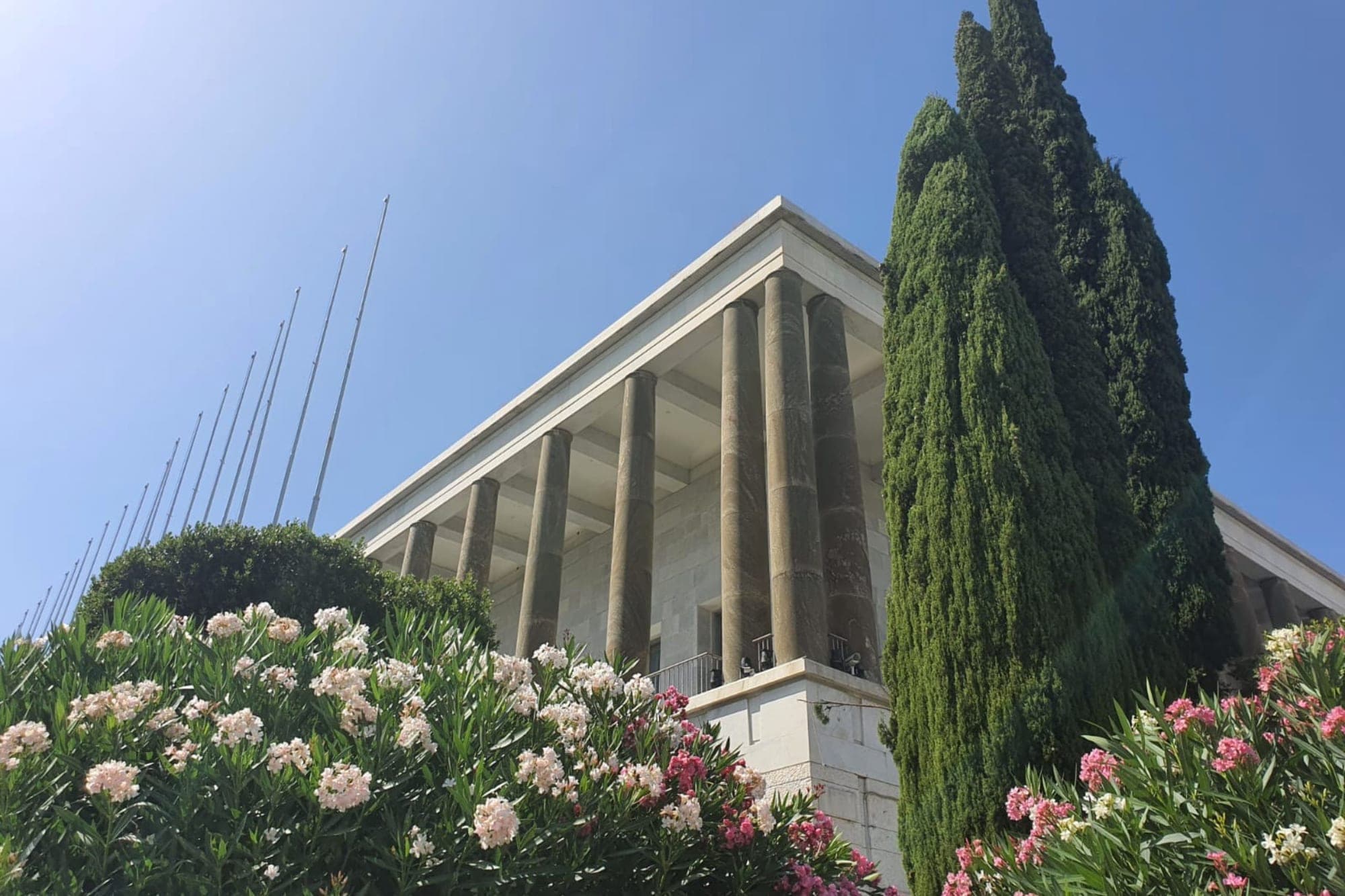 Exterior of a building with columns and a blue sky, framed by trees and flowers.