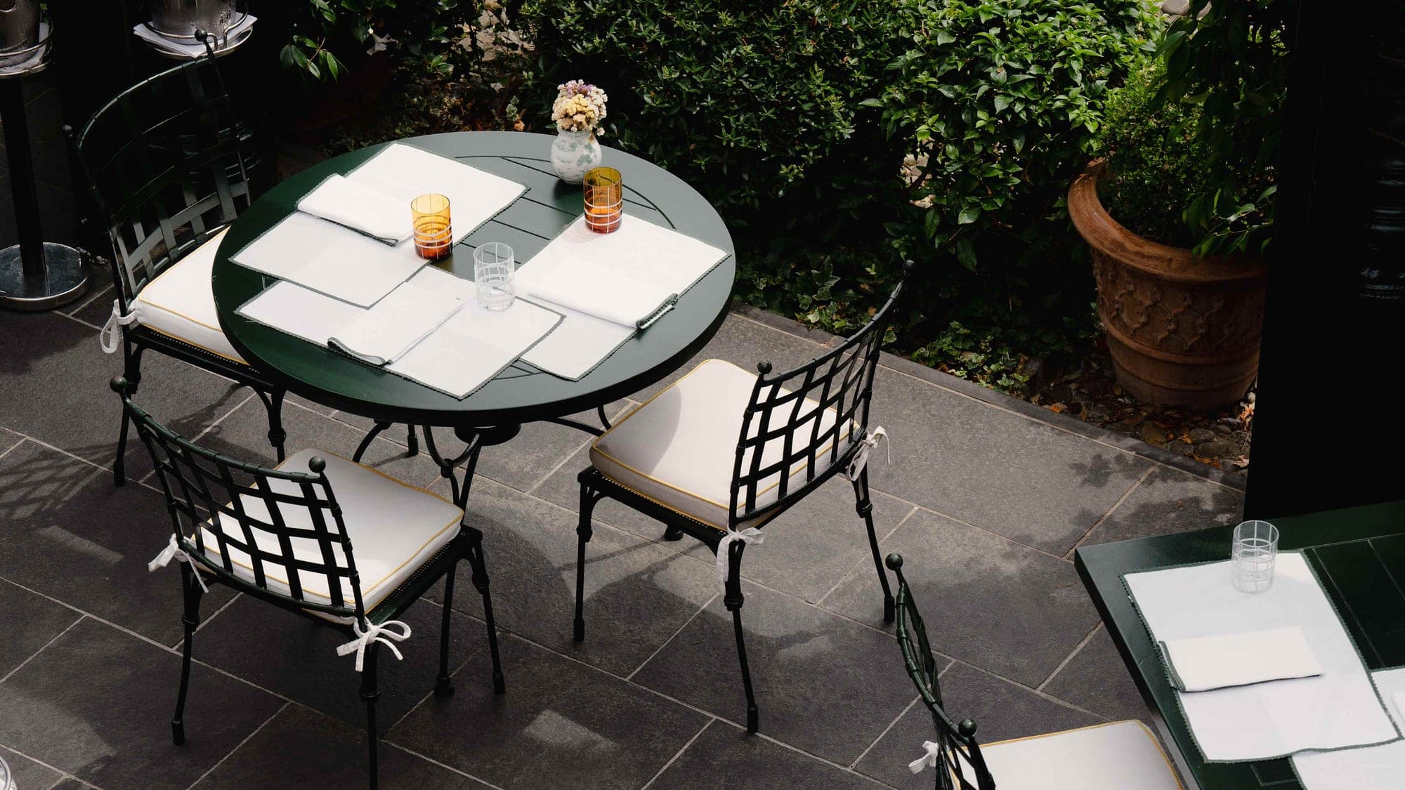 A round table set for dining on a stone terrace with chairs and greenery.