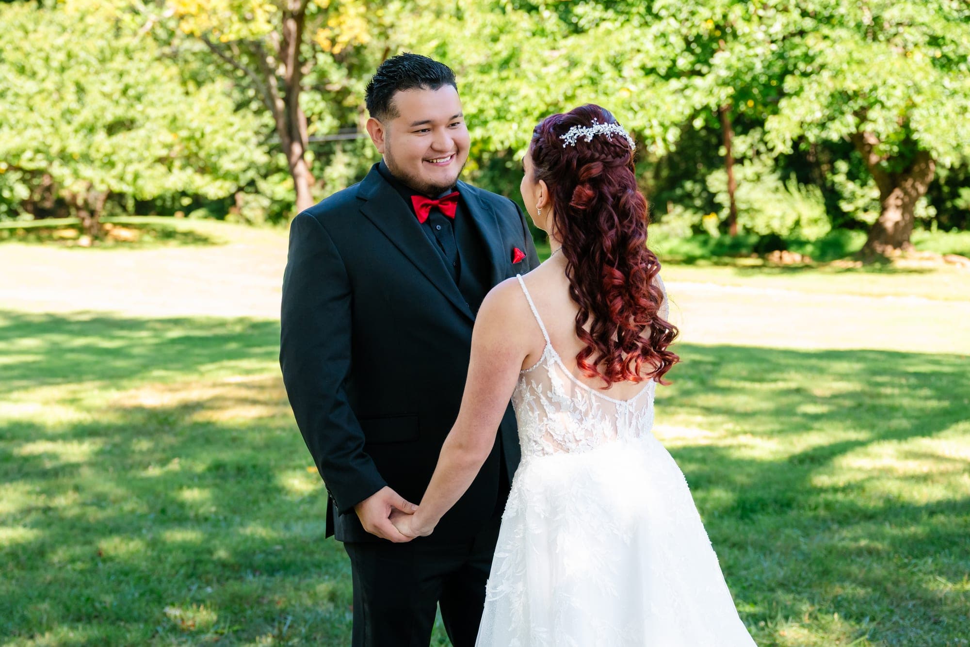 A bride and groom hold hands and smile at each other in a grassy outdoor setting.