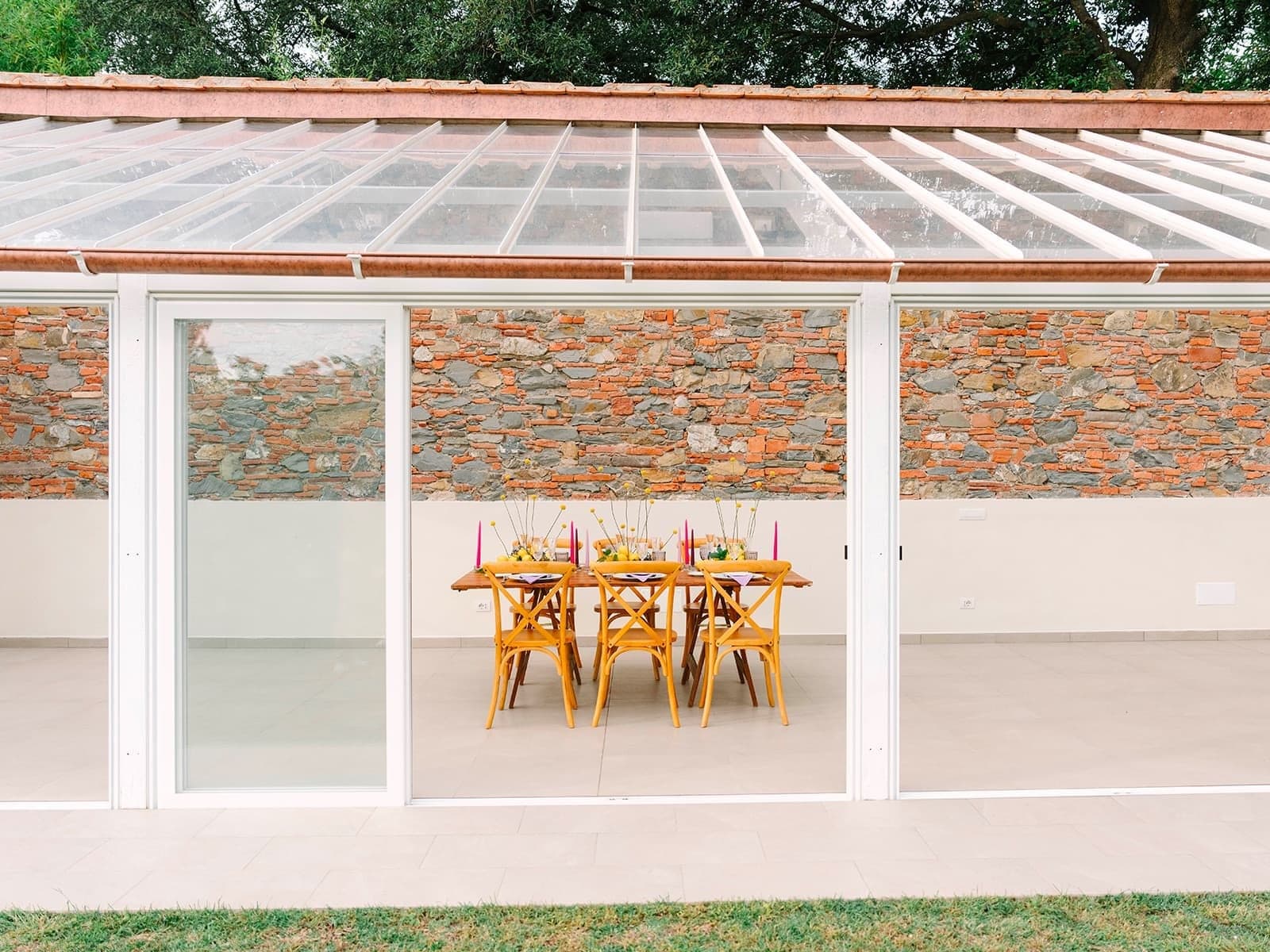 A table set for a reception inside a greenhouse with stone walls and a glass roof.