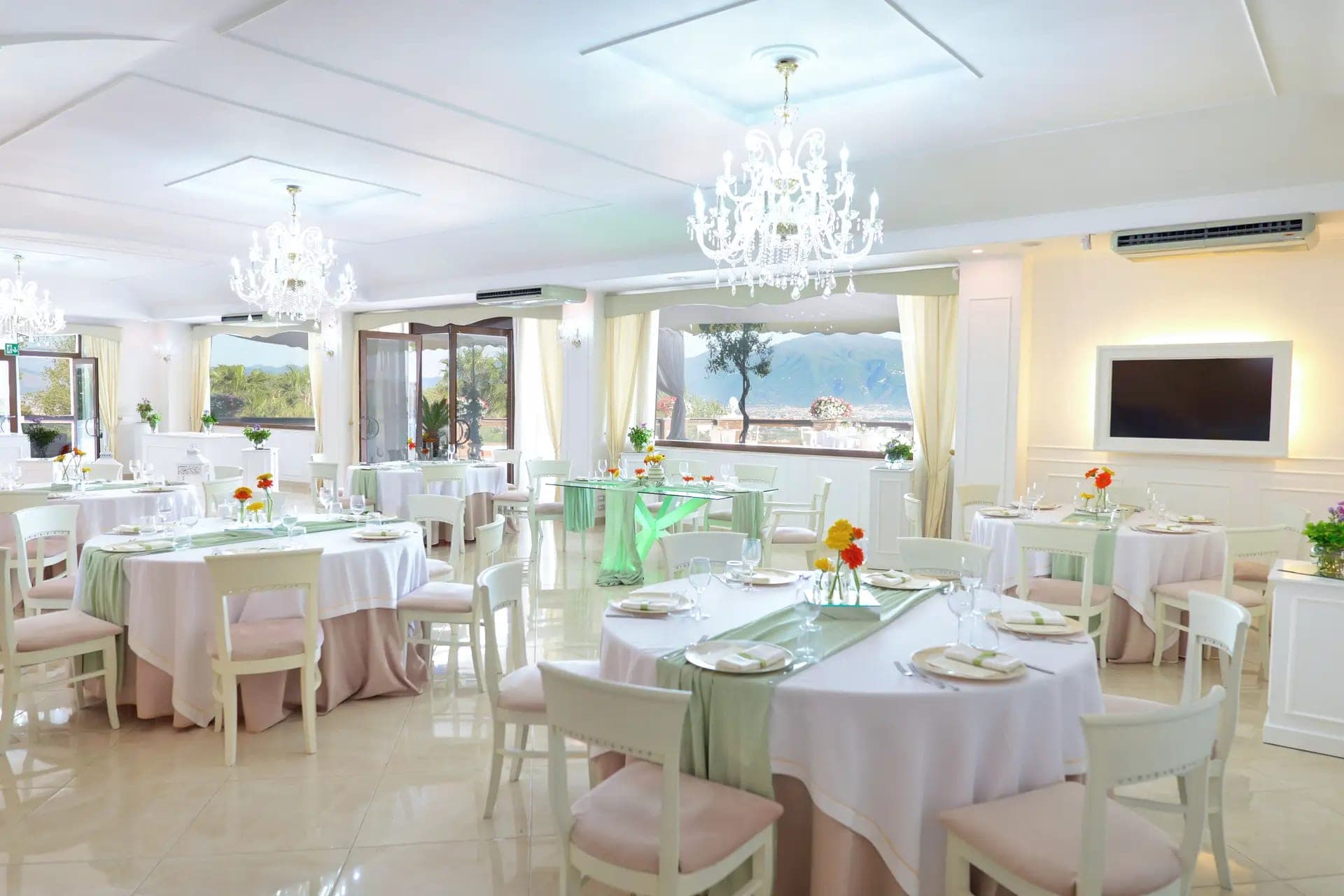 A wedding reception room with tables set for guests and a view of hills through the windows.