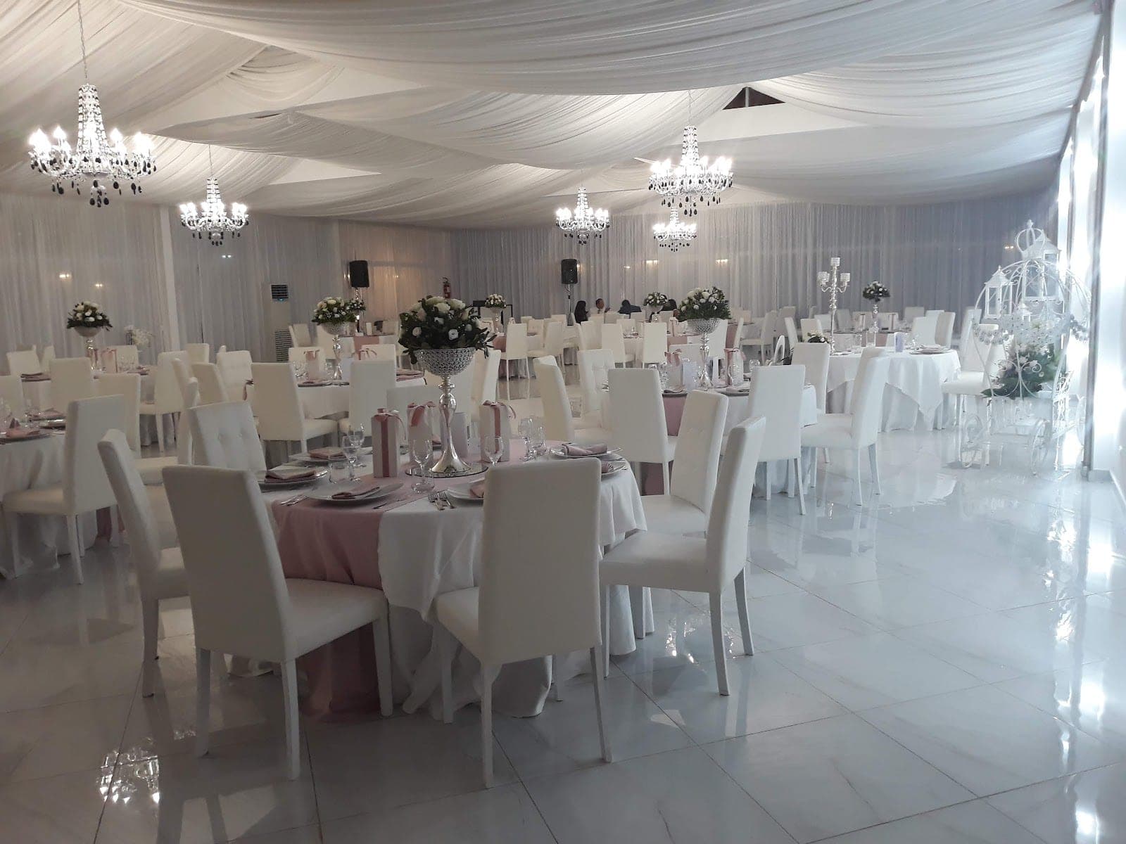 A wedding reception hall with white draped ceilings, chandeliers, and tables set for guests.