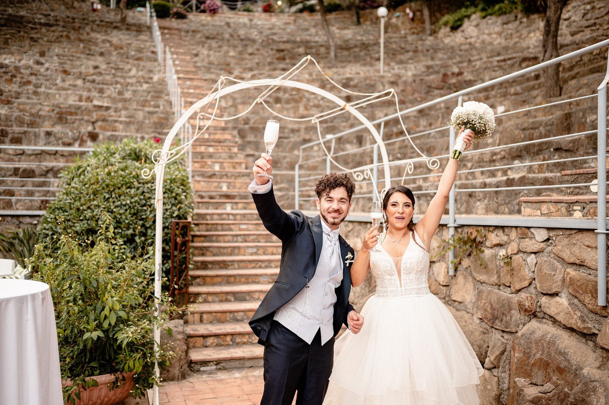 Newlyweds raise champagne glasses and a bouquet under a decorative archway.