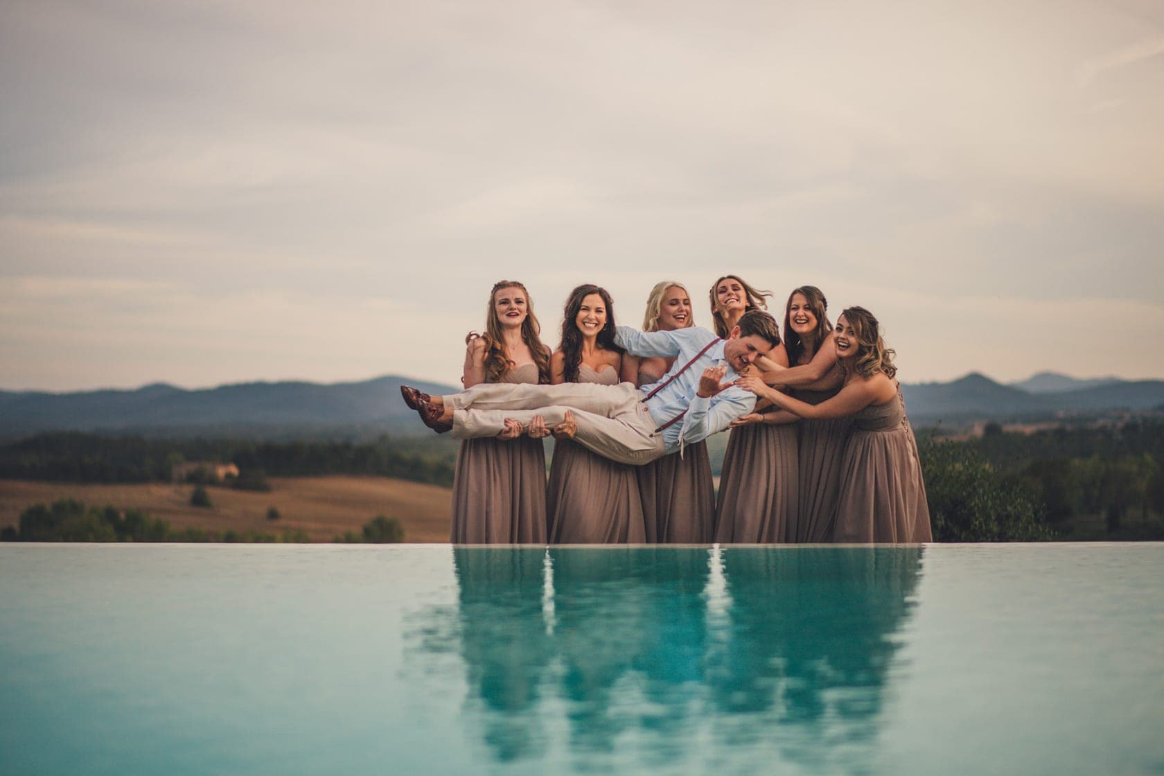 Bridesmaids holding groom over a pool with a landscape background