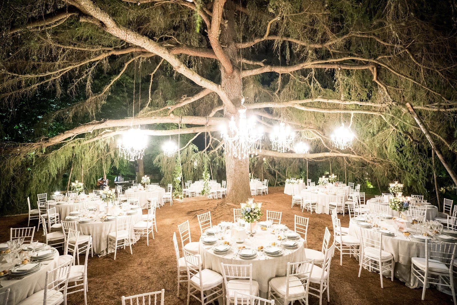 Outdoor reception tables set under a large tree with chandeliers at night.