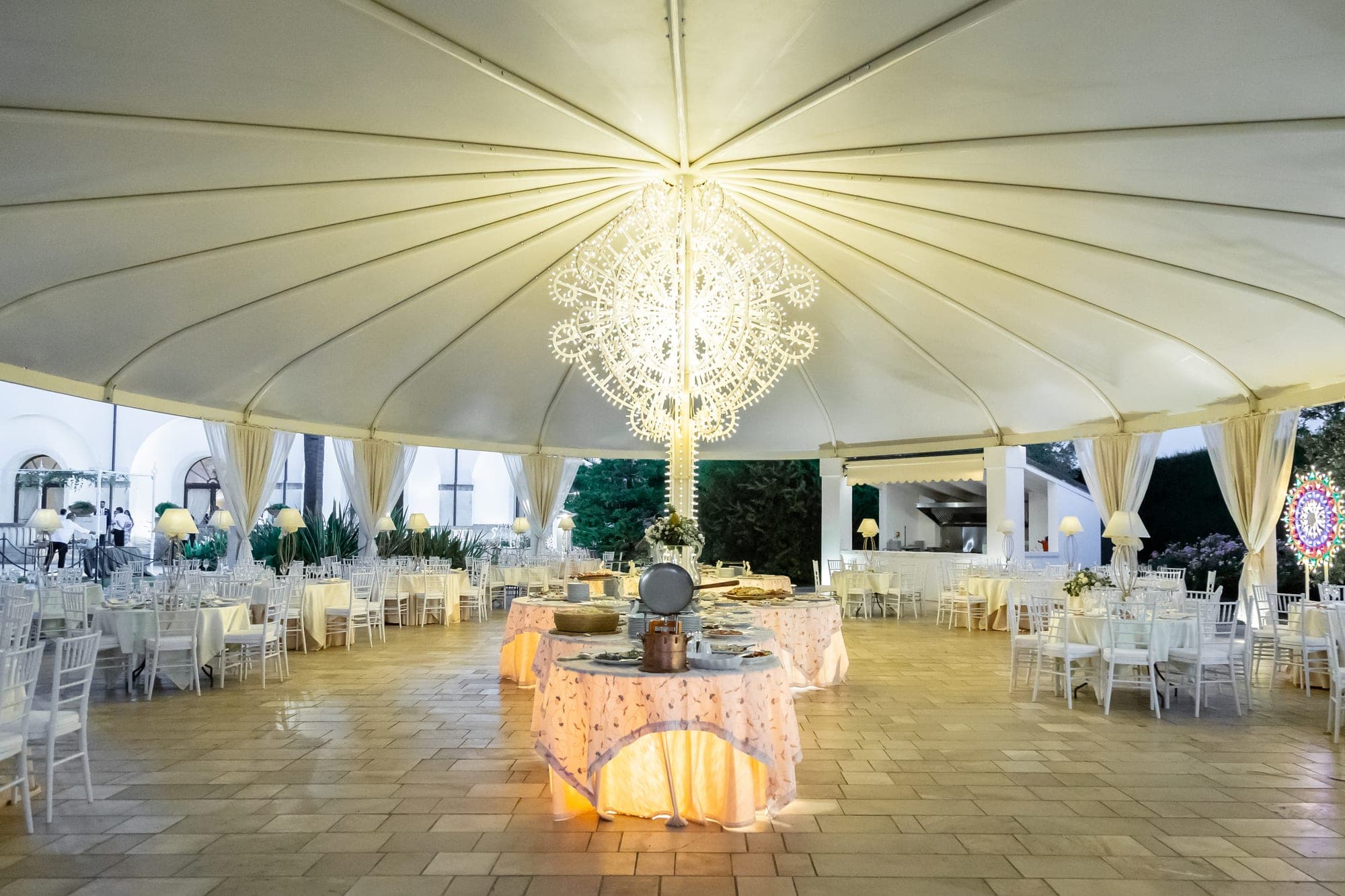 Outdoor reception area with tables set for a wedding under a large tent with a chandelier.