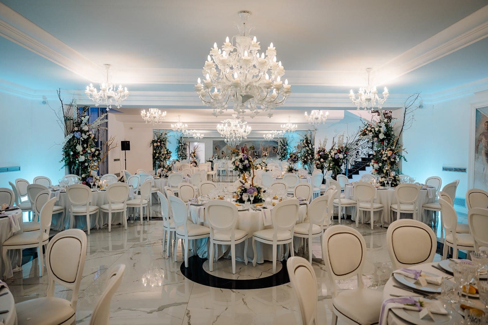 A wedding reception hall with white chairs and round tables decorated with floral centerpieces.