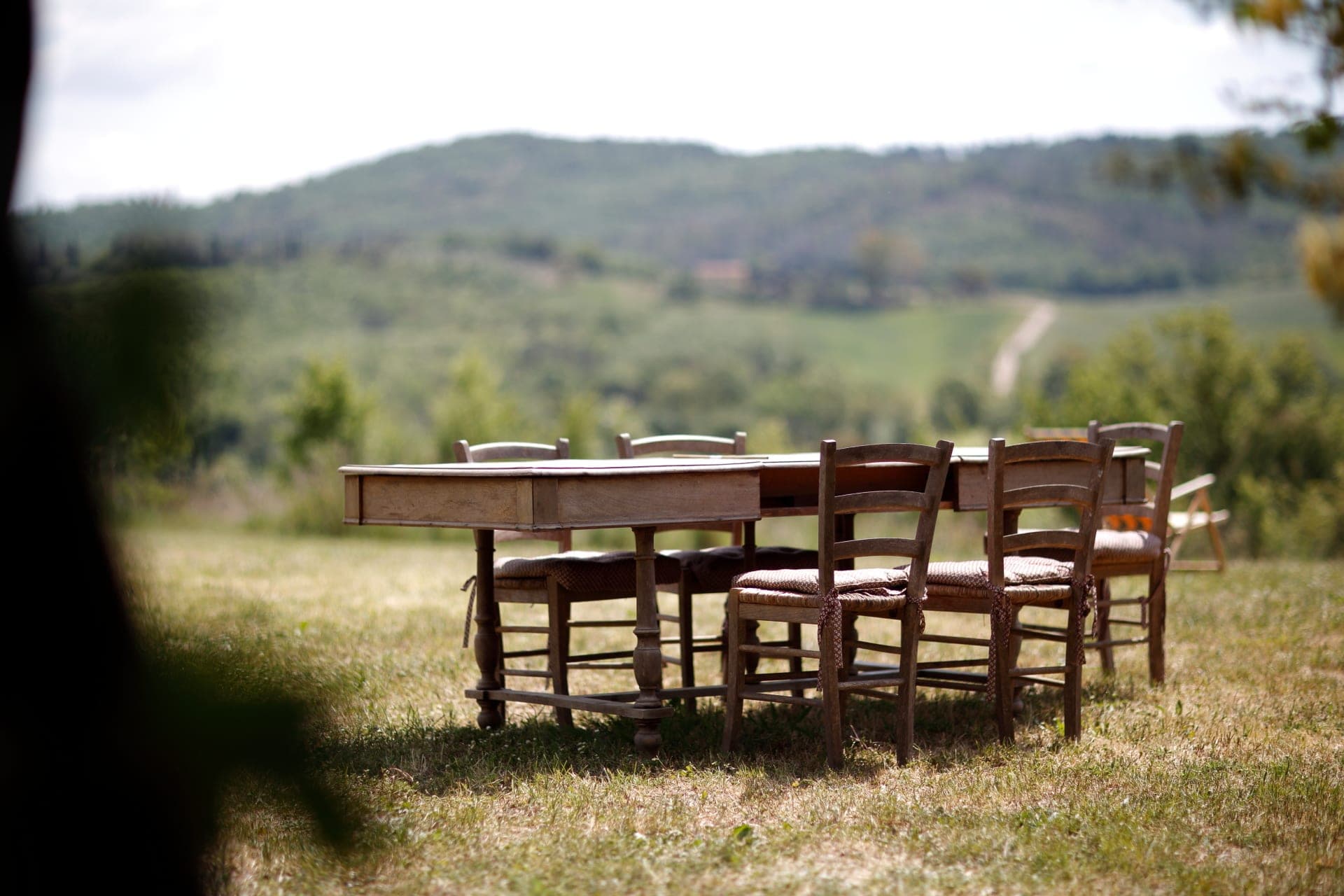 A rustic wooden table and chairs are set up on a grassy lawn with a blurred background of rolling hills.