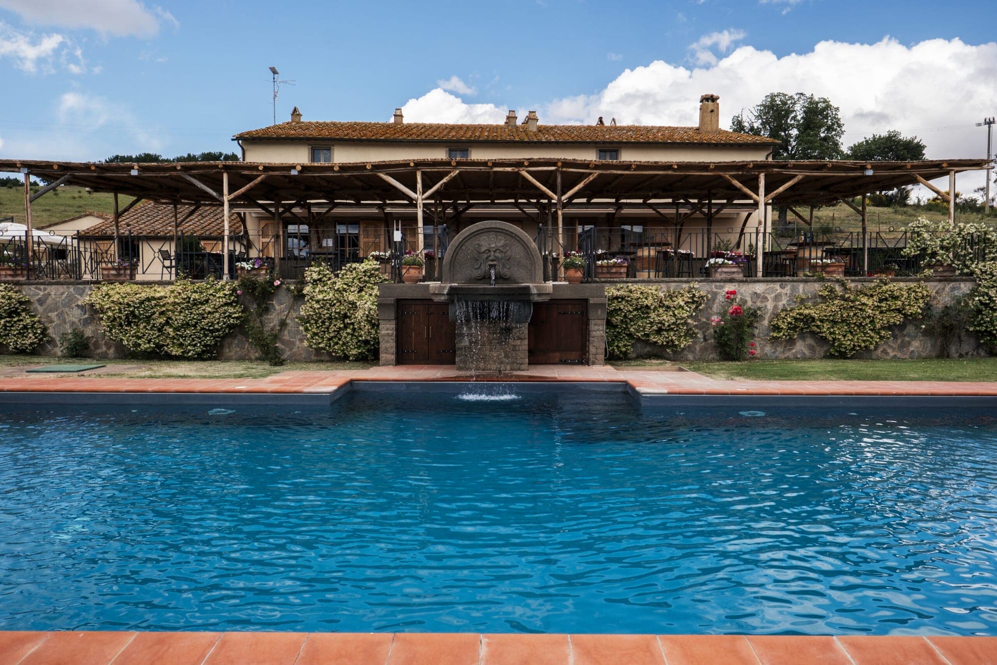 A swimming pool in front of a building with an outdoor dining area and a fountain.