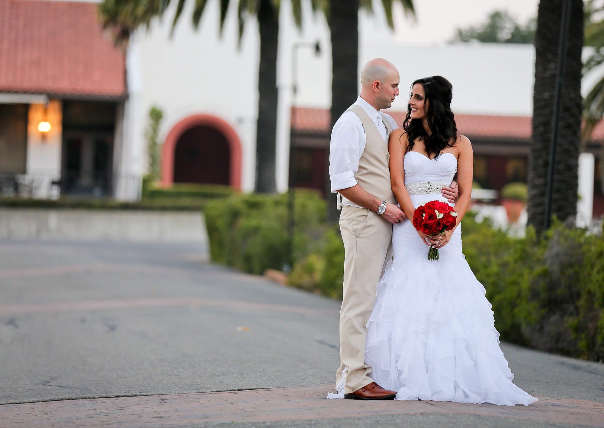 A bride and groom stand on a paved path with palm trees and a building in the background.