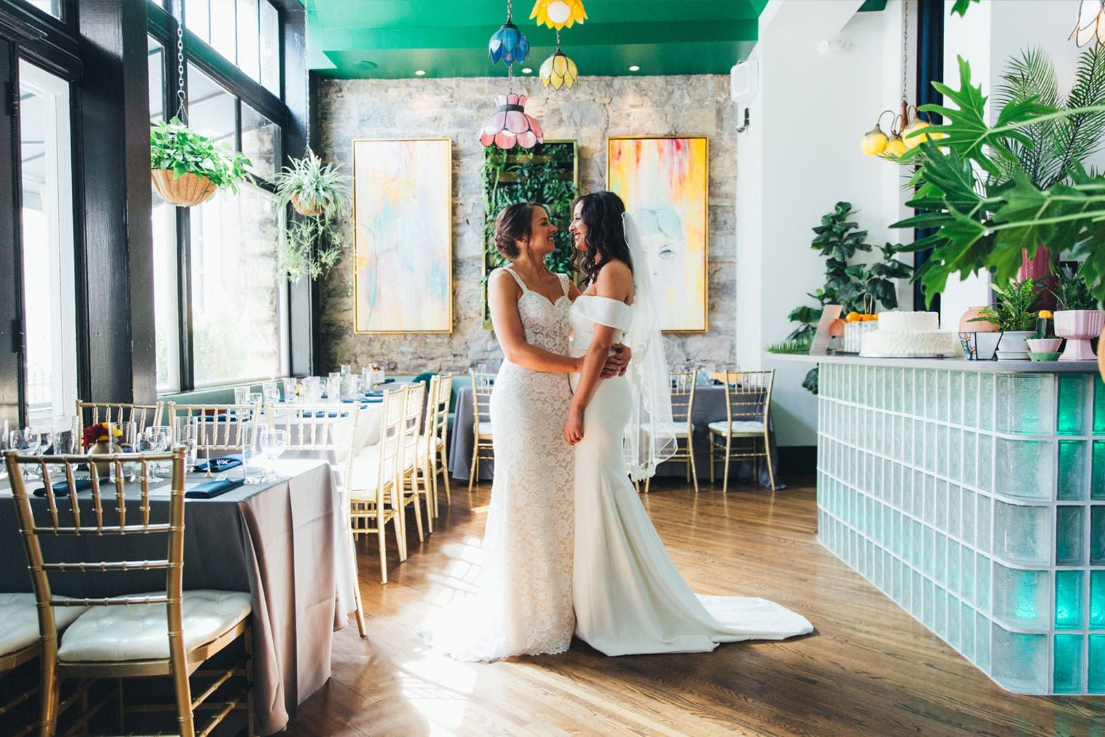 Two brides in wedding dresses embrace in a reception room with tables set for dinner.
