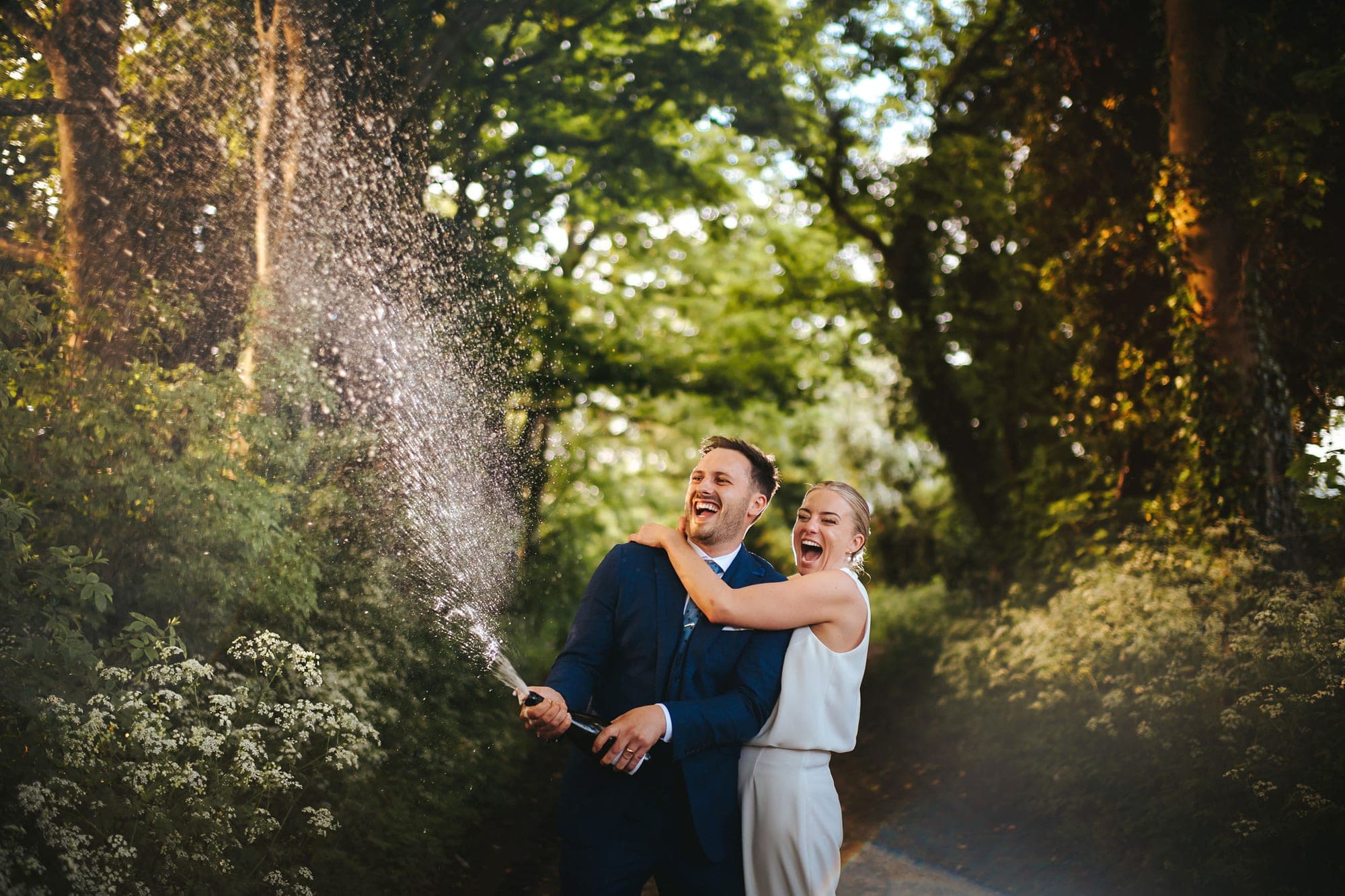 A bride and groom laugh as they spray champagne in a tree-lined outdoor area.
