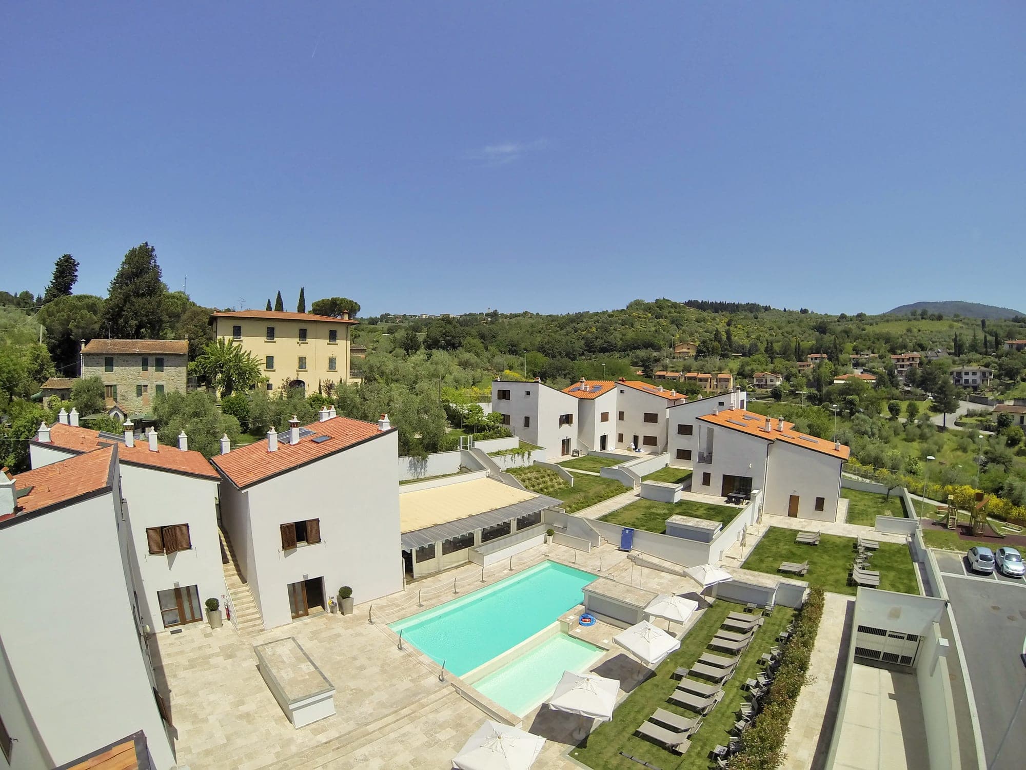 Aerial view of a resort with white buildings, a swimming pool, and green hills in the background.