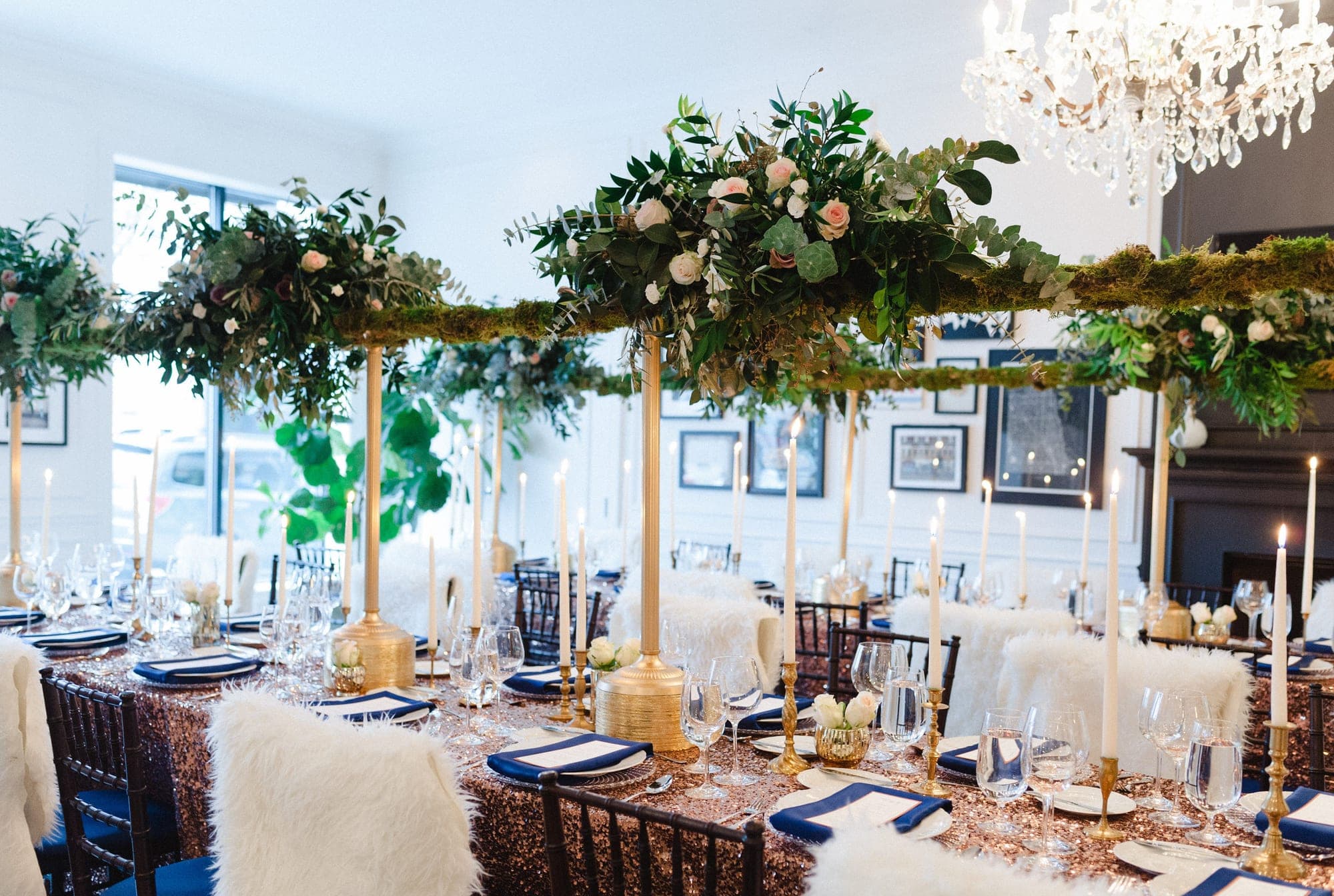 A long reception table is set with navy napkins, gold candle holders, and floral centerpieces hanging overhead.