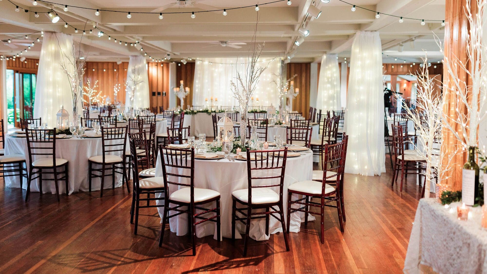 A wedding reception hall with round tables, chairs, and decorative lighting.