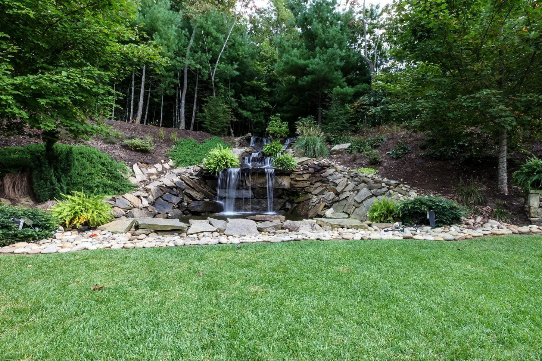 A tiered waterfall cascades over rocks into a pool, surrounded by green grass and trees.