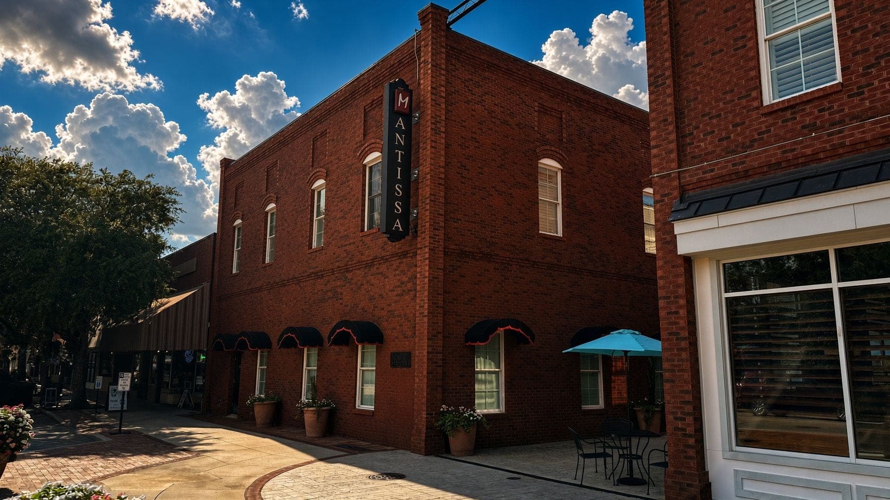 Exterior of a brick building with a vertical MANTISSA sign and a blue umbrella outside.
