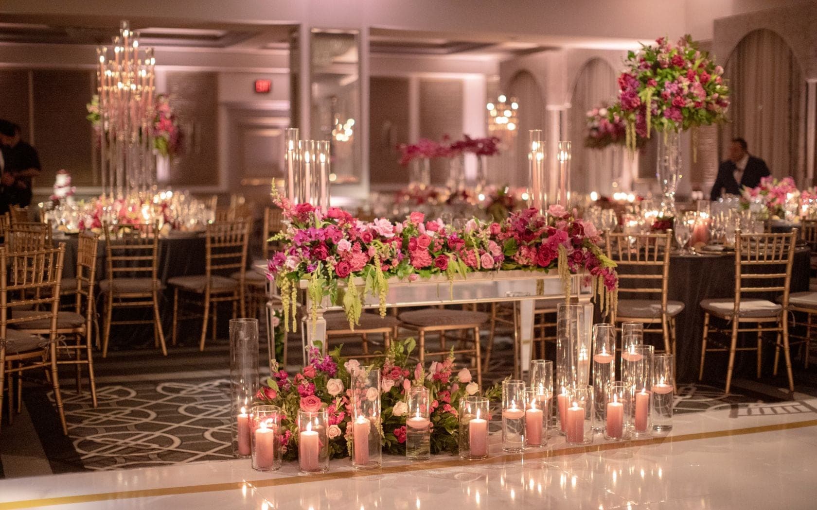 A wedding reception hall with tables set for dinner, adorned with pink floral arrangements and lit candles.