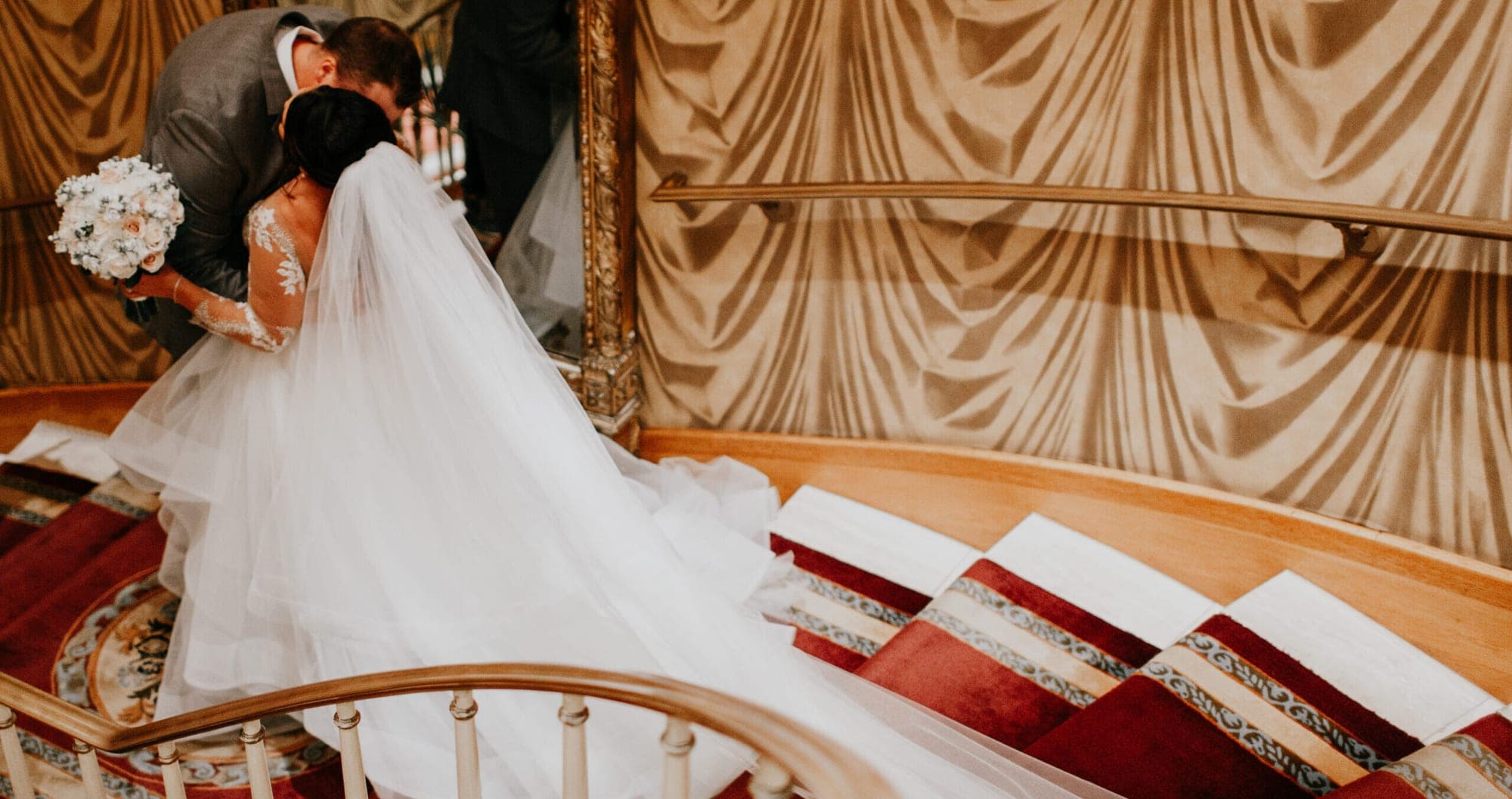 A bride and groom kiss on a carpeted staircase with ornate wall coverings.