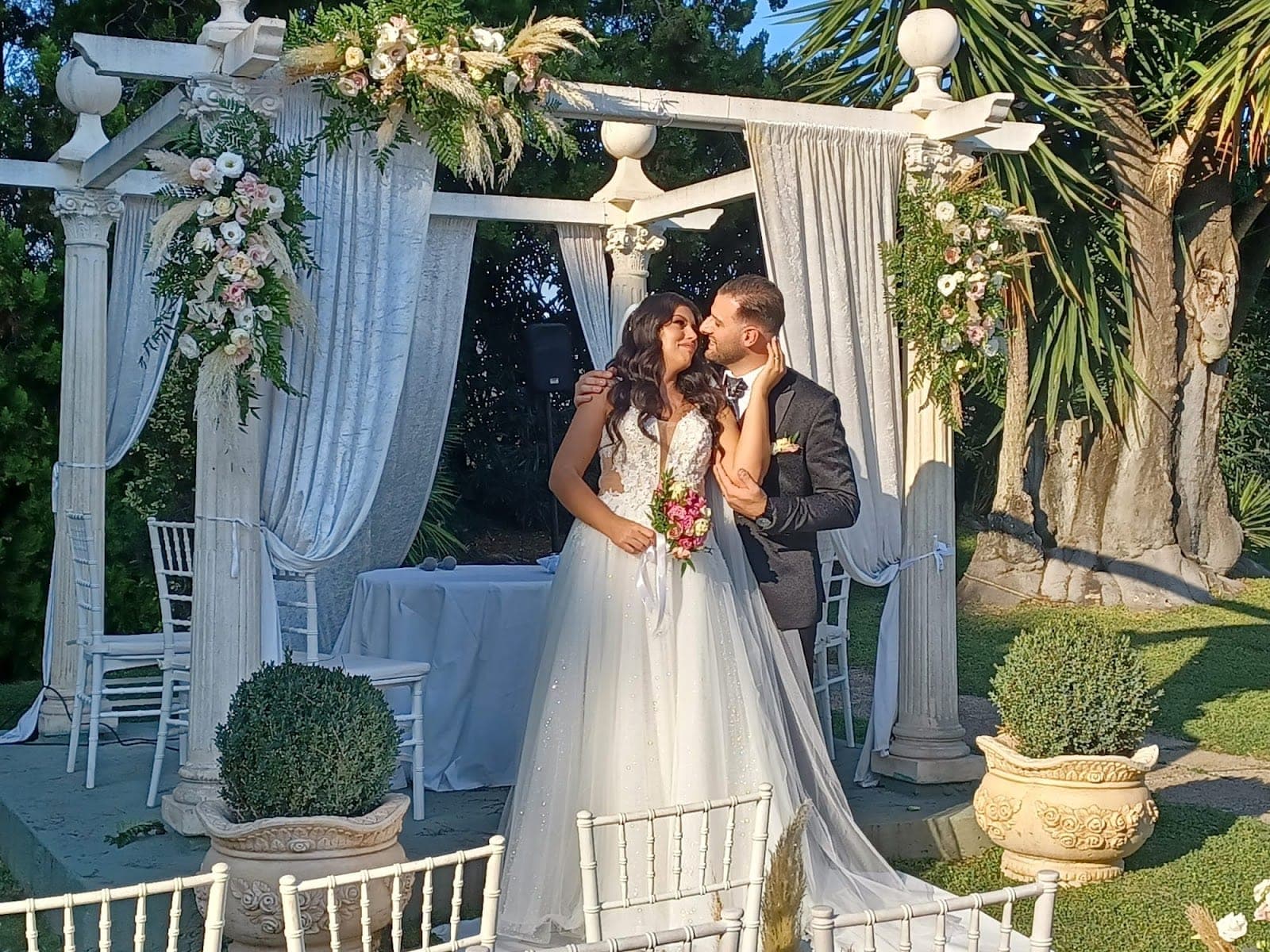 A bride and groom embrace in front of a white wedding arbor decorated with flowers.
