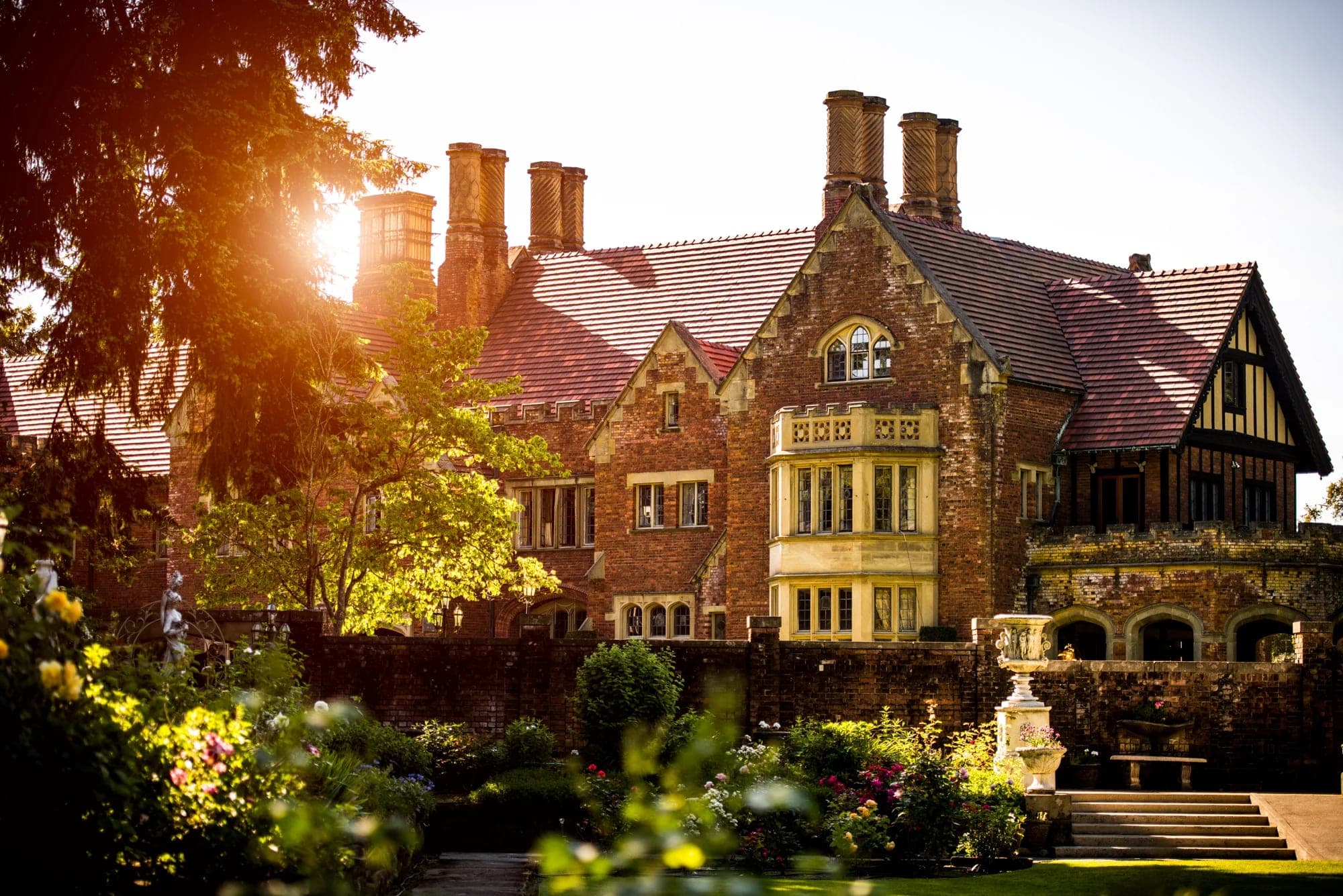 A brick castle with multiple chimneys and a bay window, seen through trees and a garden.