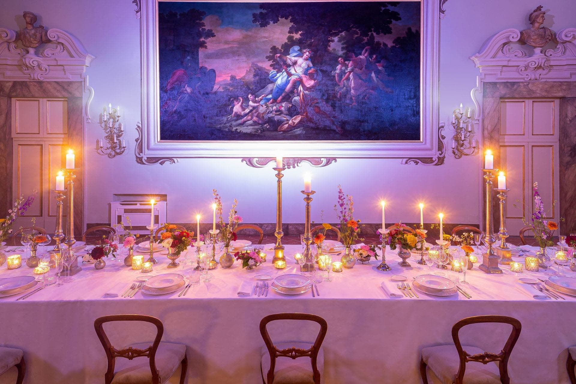 A long dining table set with white linens, floral centerpieces, and candelabras under a large painting.