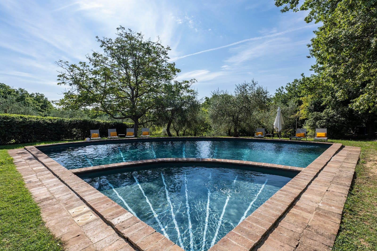 A swimming pool with brick edging and loungers under trees.