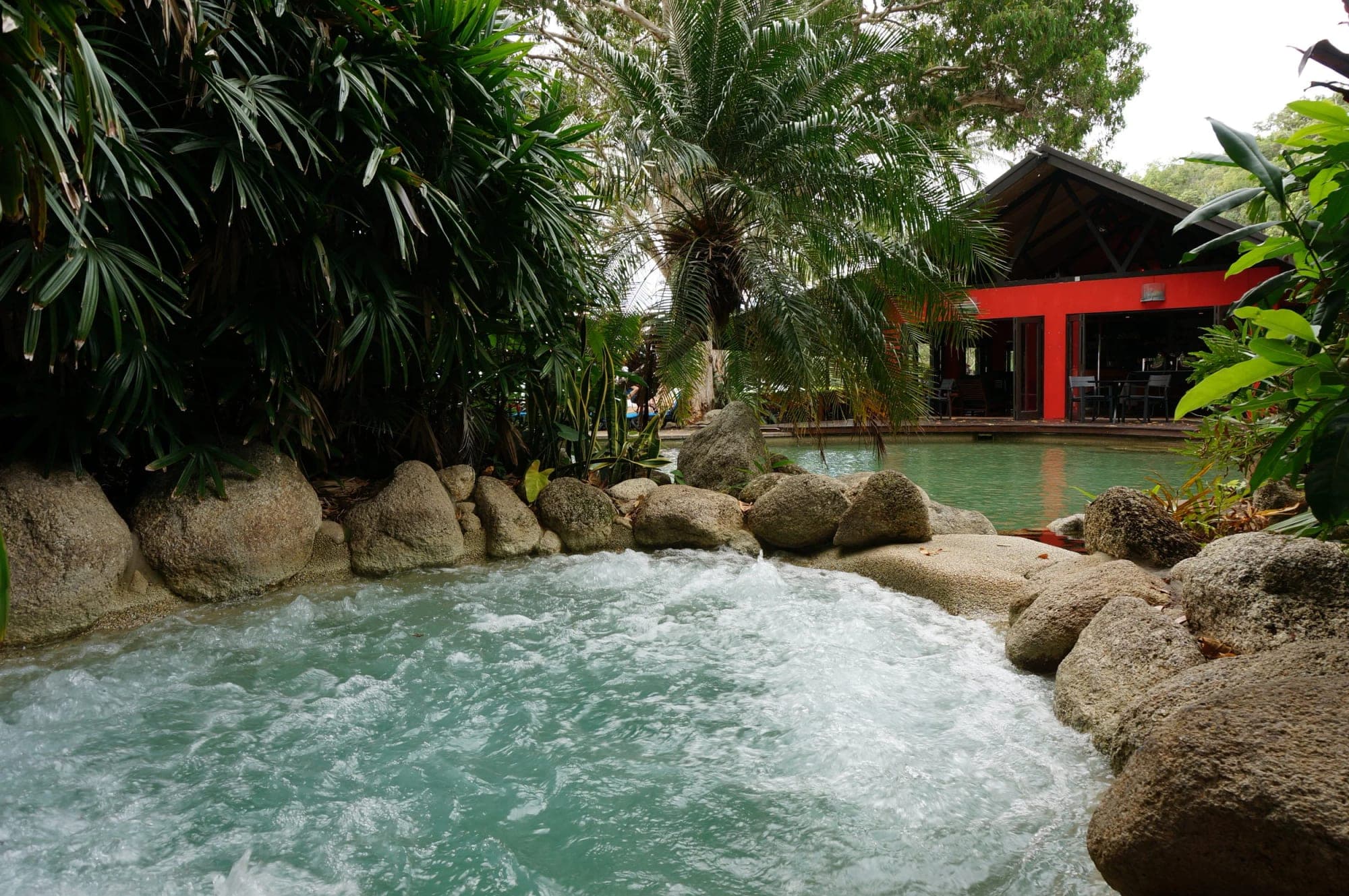 A bubbling pool with rocks and lush greenery, leading to a red building with tables and chairs.