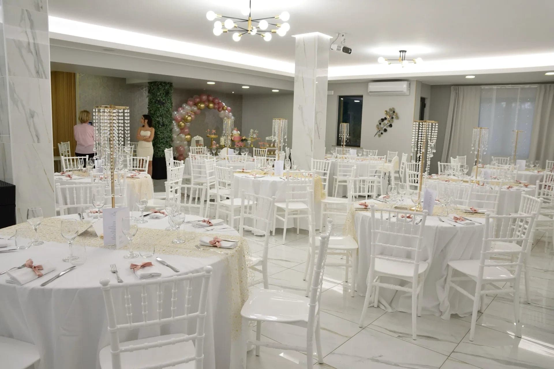 A reception hall with round tables set for a wedding, white chairs, and floral decorations.