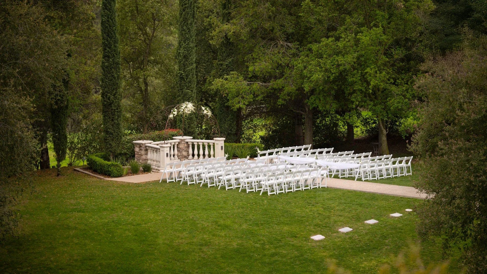 Outdoor wedding ceremony setup with white chairs on a green lawn and trees in the background.