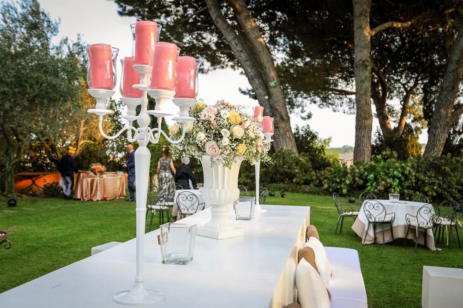 A garden reception setup with a long white table, floral arrangements, and trees in the background.