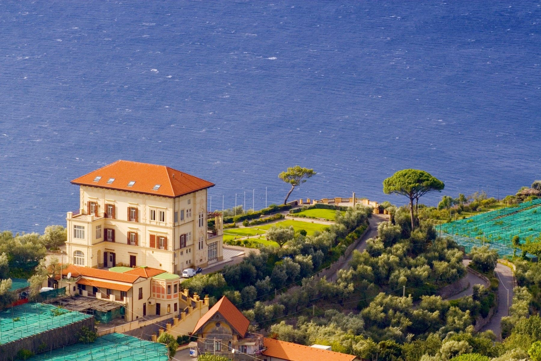 A villa with an orange roof sits on a hillside overlooking the blue sea.