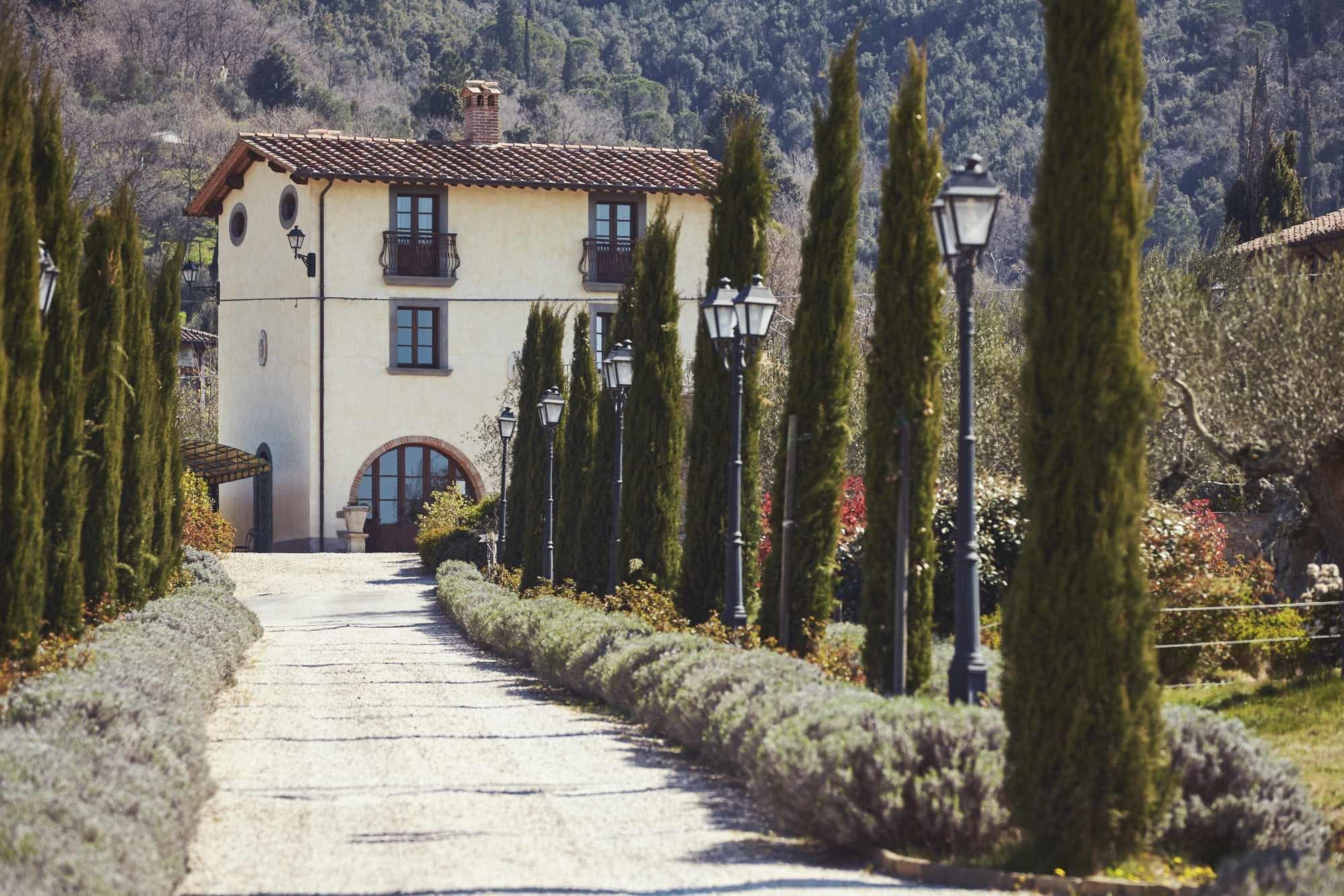 Driveway leading to a villa with cypress trees and lampposts.