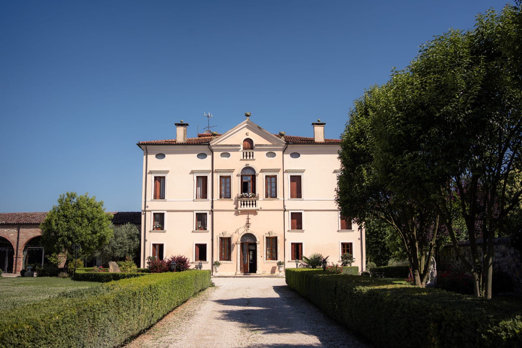 Exterior of a pale yellow villa with a central balcony and arched doorway, surrounded by manicured gardens and trees under a clear blue sky.