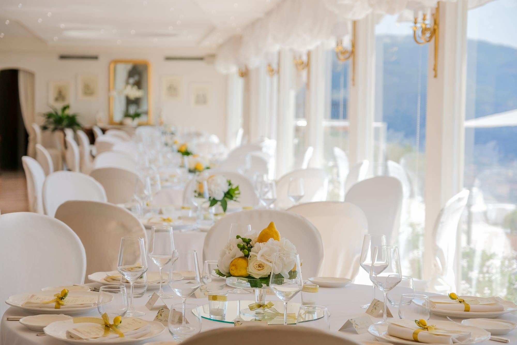 A long reception table is set with white linens, plates, glasses, and floral centerpieces.