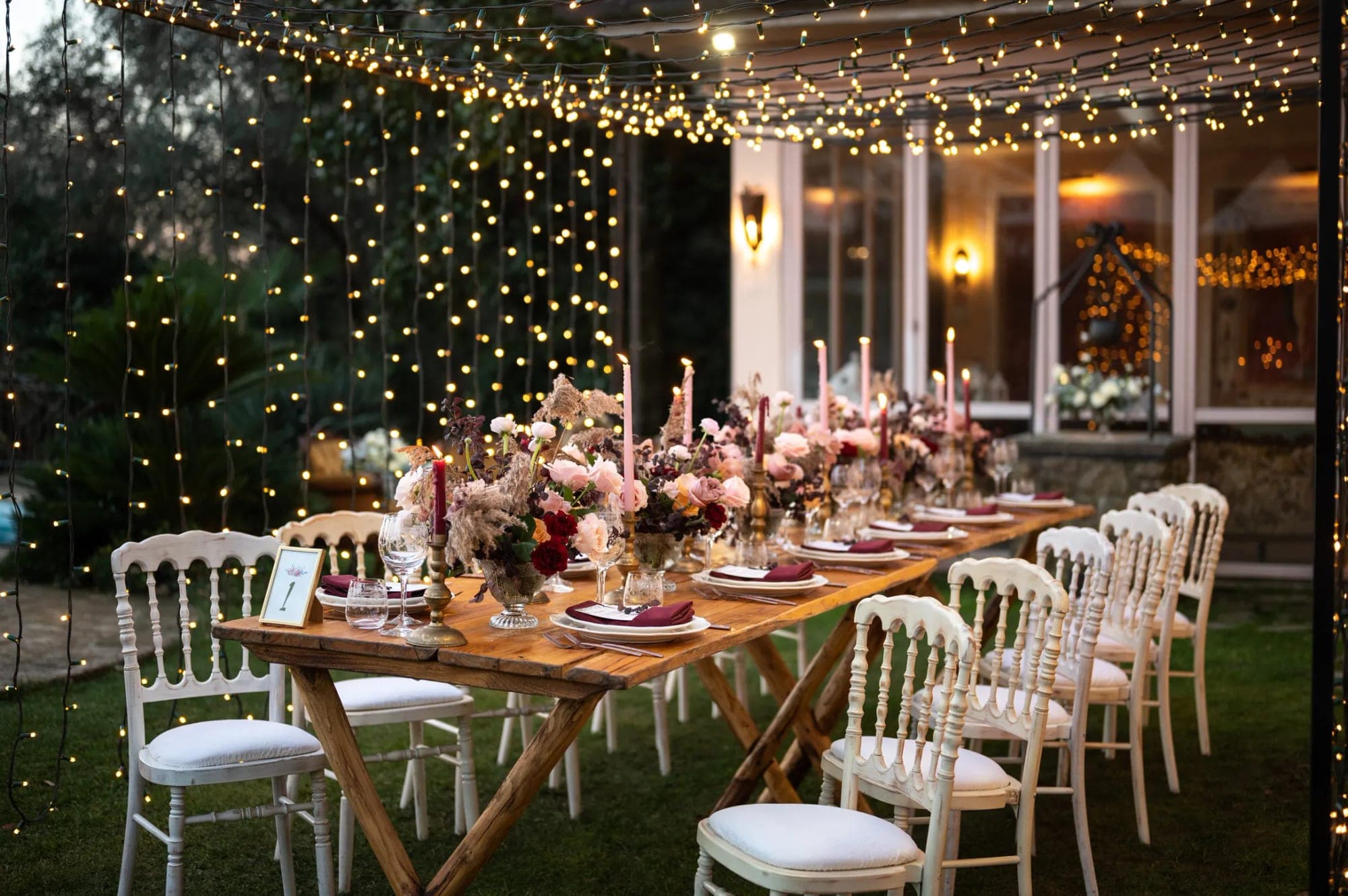 Outdoor wedding reception dining table set with floral arrangements and fairy lights.