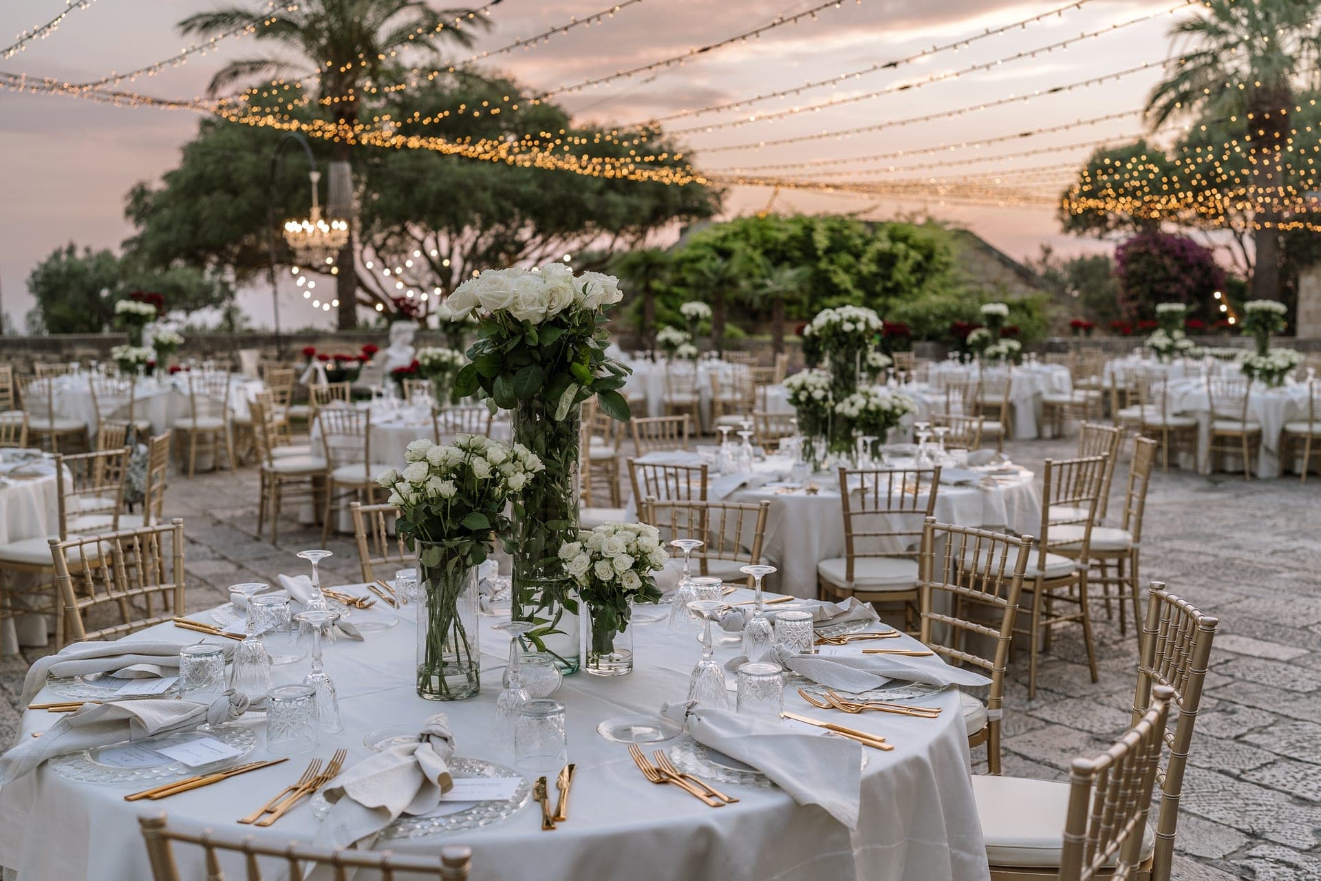 Outdoor reception tables set with white linens and floral arrangements under string lights.